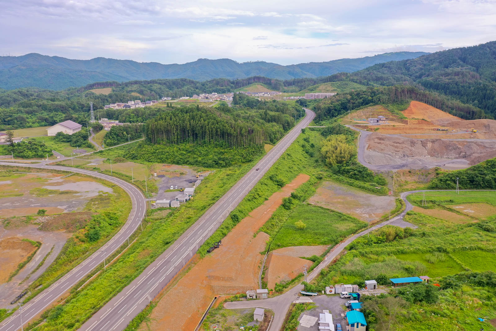 中央を高速道路が通り、周囲には緑の丘や森林が広がり、ところどころに開けた土地や小さな建物が点在する空から見た田舎の風景の写真