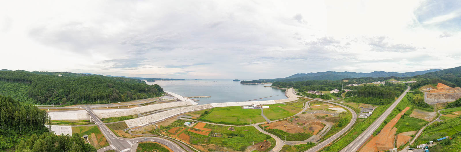 中央に海が広がり、その周囲を緑の丘や森林が囲み、海岸にはコンクリートの防波堤や道路が整備され、一部には工事中の土や重機が見える曇り空の下の沿岸地域の写真