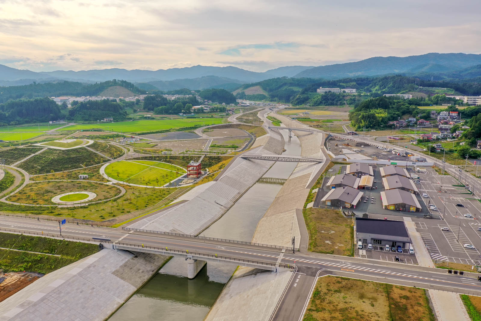 川にはコンクリートの護岸が施され、複数の橋が架かり、周囲には緑の畑や道路、車の停まった駐車場、建物が点在し、背景には丘や山が広がる空から見た風景の写真