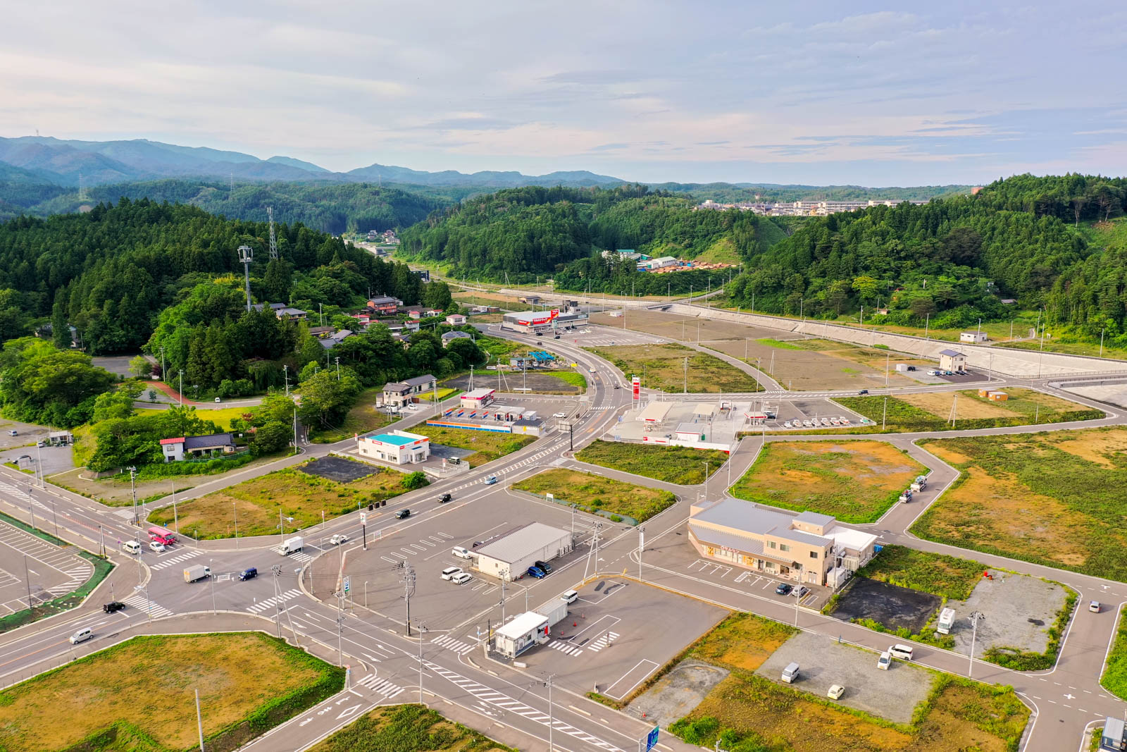 緑豊かな丘や森林に囲まれた小さな町を空から見下ろした風景で、建物や道路、駐車場、草地が点在し、背景には山々が広がる曇り空の下の写真