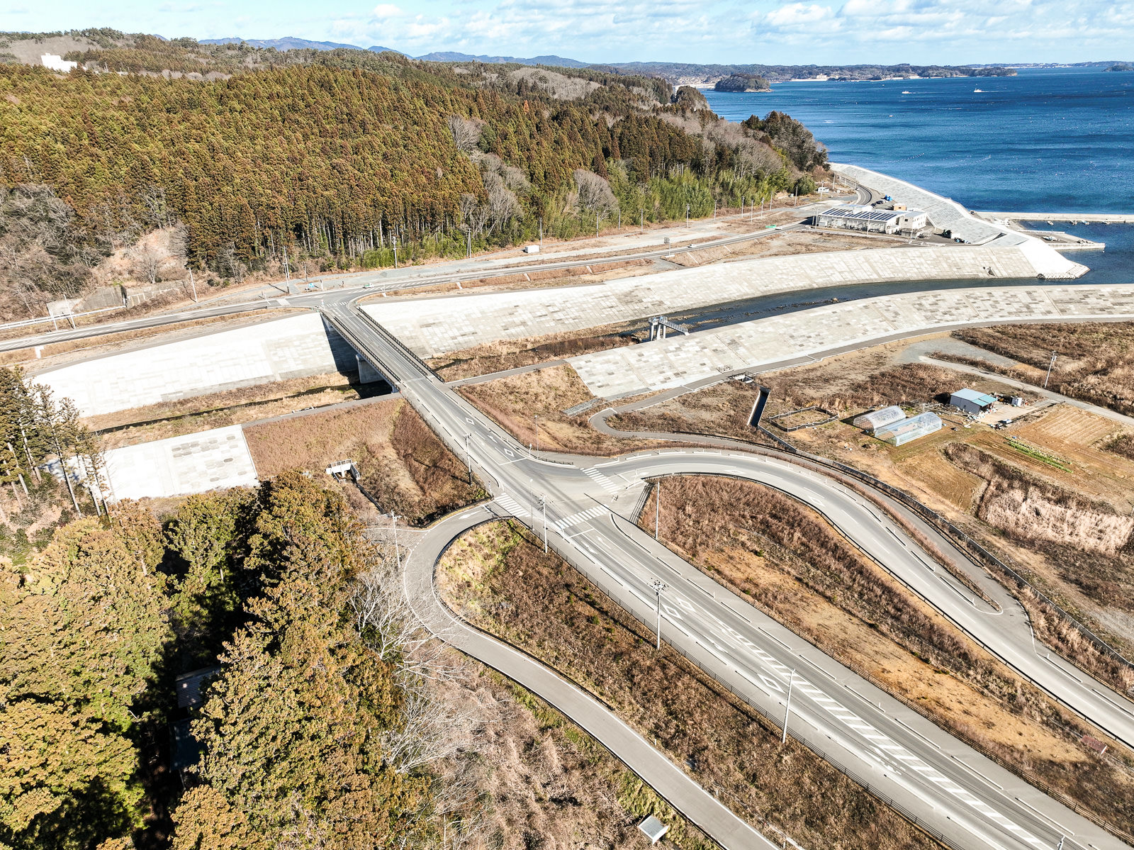 海に近いエリアで山林と海の間に長く延びた防潮堤と交差する道路が確認でき、全体的に復興工事中の様子がうかがえる昼間の風景を高台から定点カメラで撮影した写真