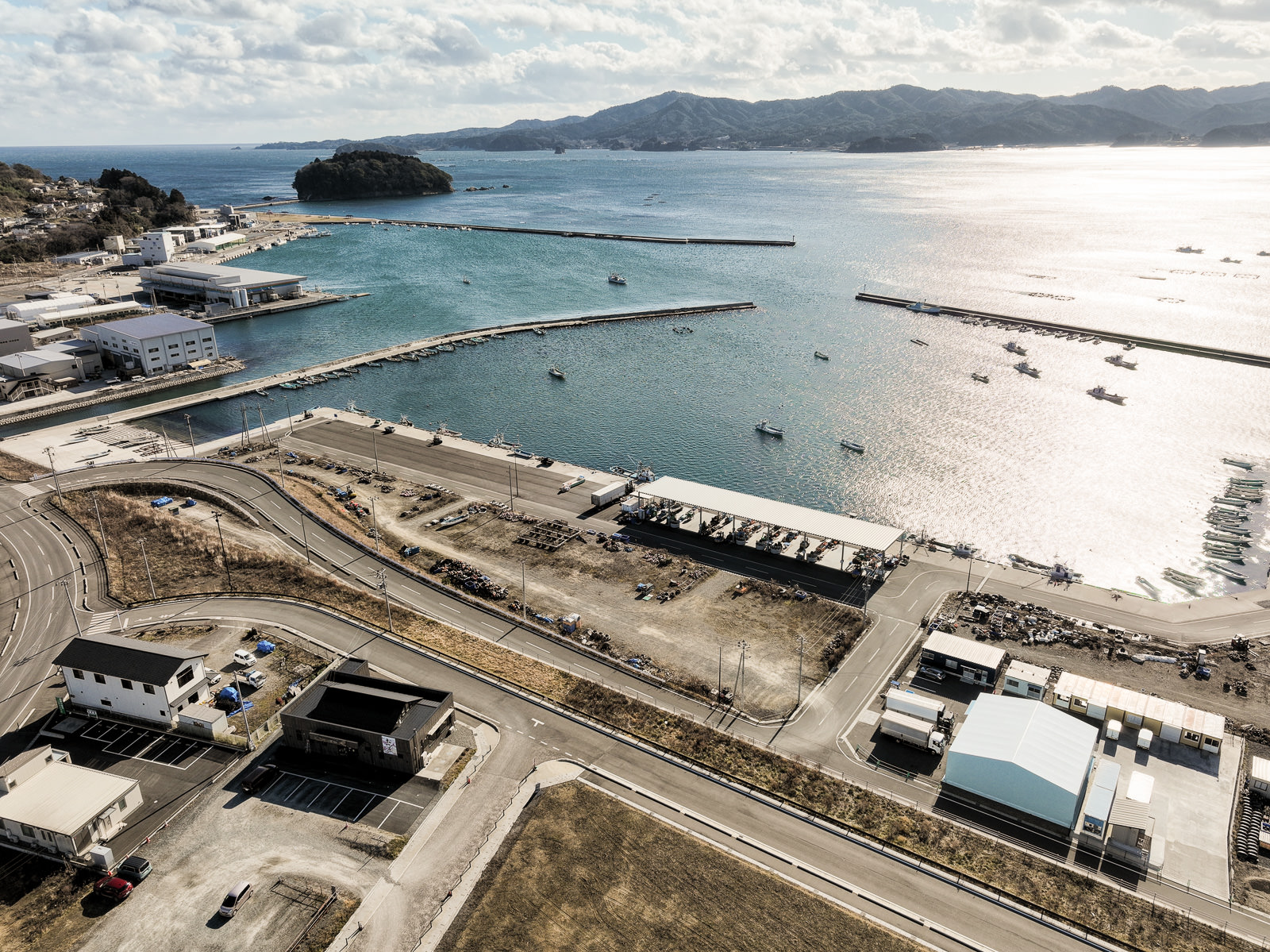 防波堤内に漁船が浮かび、岸には白い倉庫や工場が整然と並んでいる海に面した漁港の風景を高台から定点カメラで撮影した写真