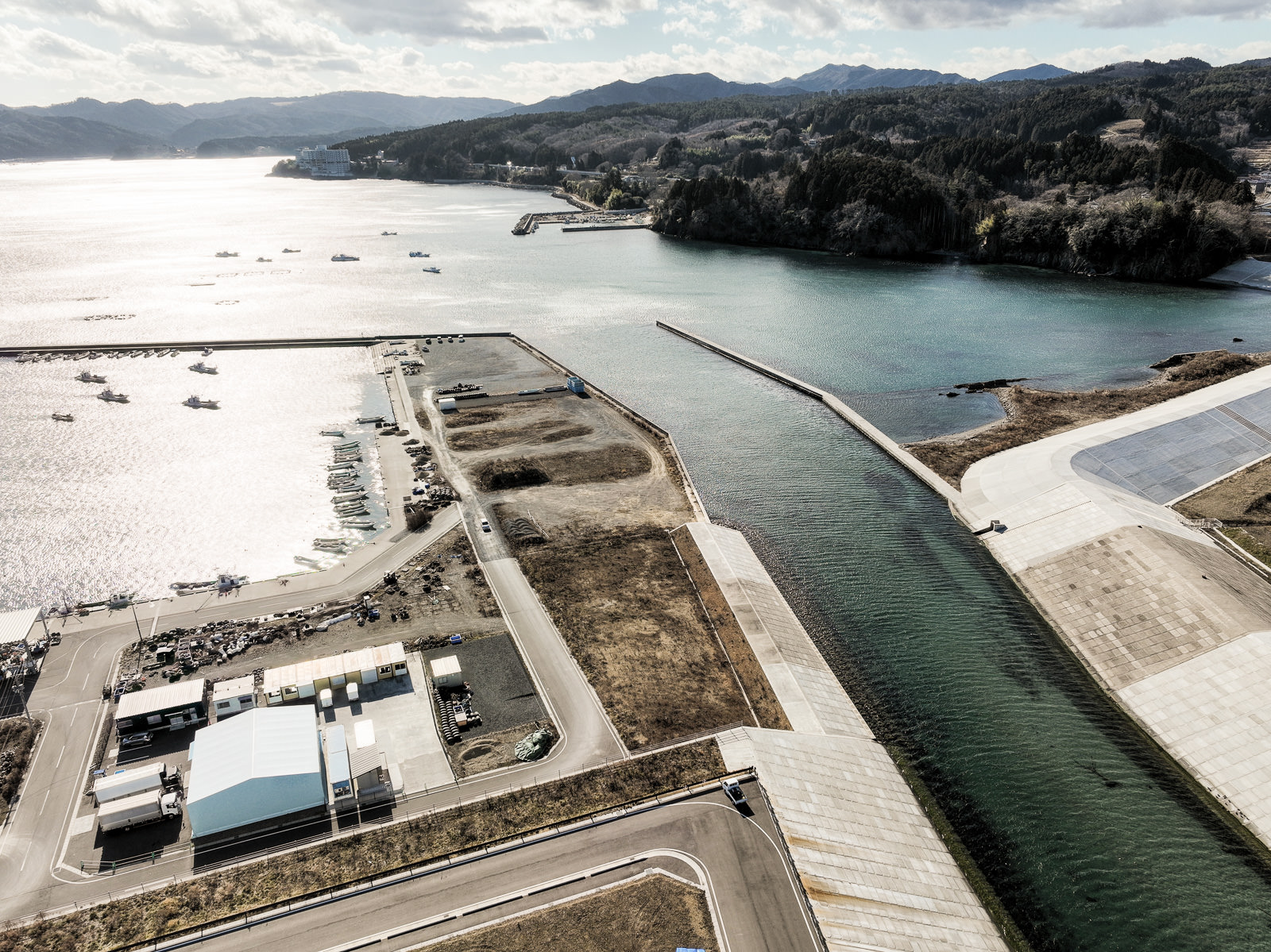 防波堤に囲まれた入り江の奥に、静かに停泊する漁船と整備された埠頭があり、背景には山と森が広がっている風景を高台から定点カメラで撮影した写真