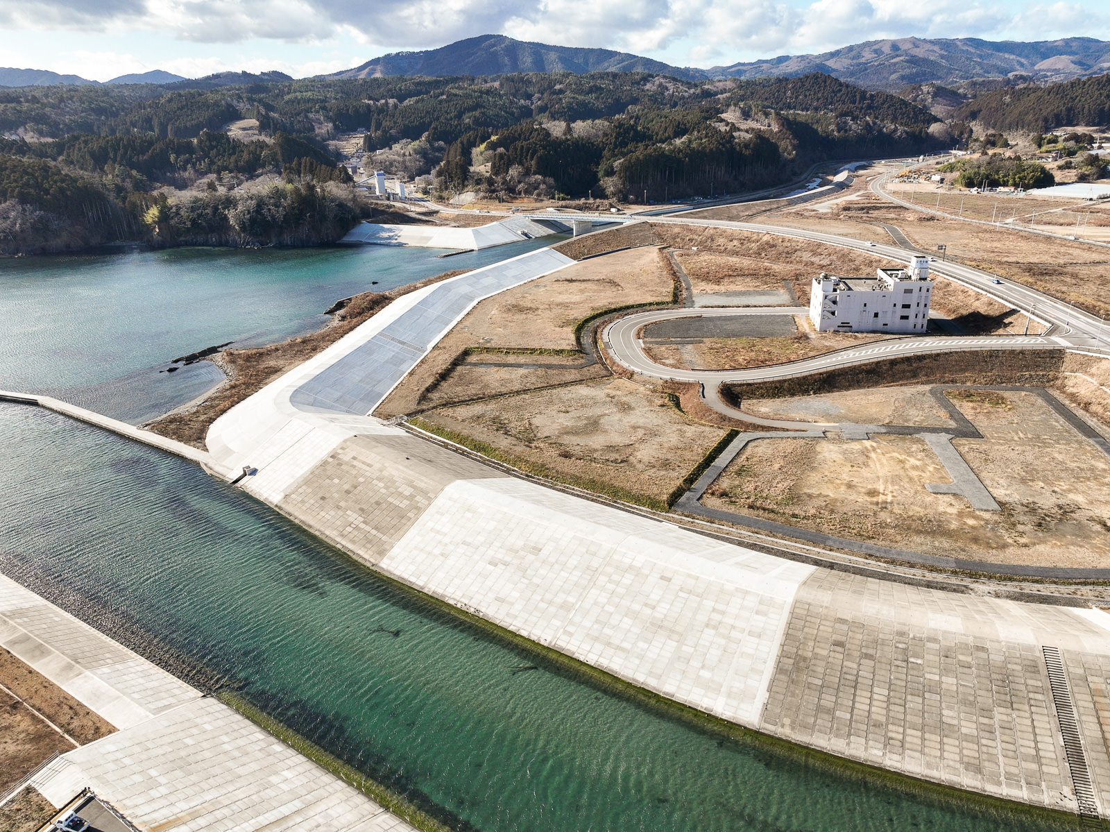 海へ流れ込む川の両岸に高さのある堤防が築かれ、その奥には曲線を描く道路と、白い避難ビルのような建物が広がる造成地とともに、山に囲まれた静かな風景を高台から定点カメラで撮影した写真