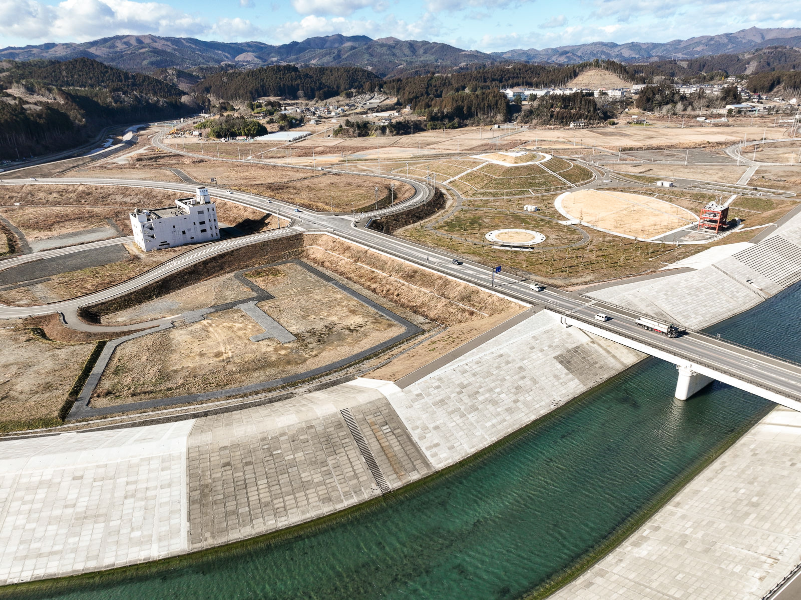 川の上にかかる大きな橋を中心に、その奥には円形の公園や広場、赤い建造物が点在し、さらに背後には山々が広がる整備された土地が一望できる風景を高台から定点カメラで撮影した写真