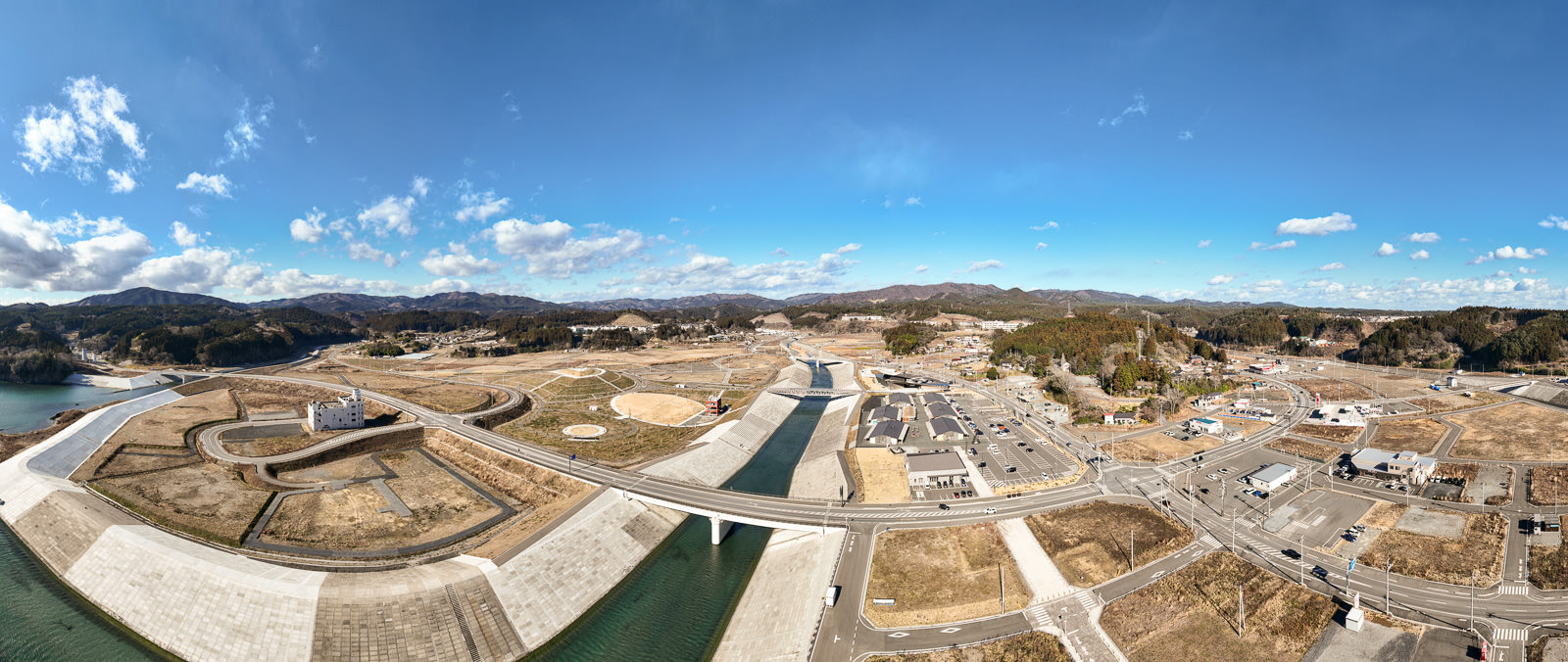 手前に川と防潮堤、中央に大きな橋と新しく整備された道路網、その奥に広がる山々と街並みがパノラマで写されており、復興が進んだ都市計画の昼間の風景を高台から定点カメラで撮影した写真