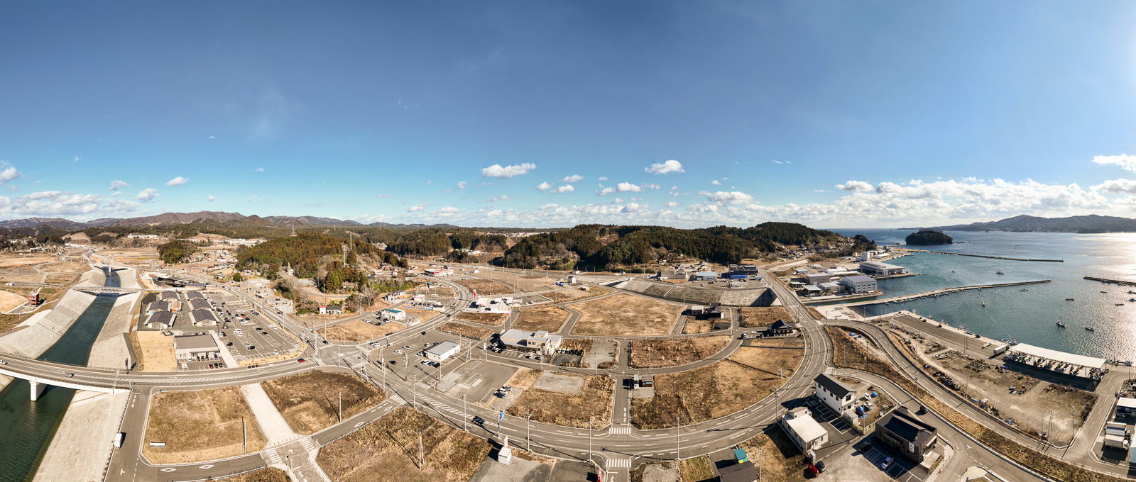 手前から奥へと川と橋が続き、右手には広く整備された街並みと道路網、さらにその先に港と海が広がっており、港湾地域の再整備の様子の昼間の風景を高台から定点カメラで撮影した写真