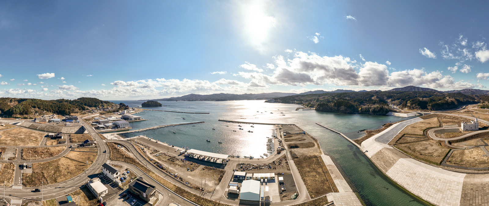 中央に太陽が輝き、左右に湾と堤防、周囲に山々と整備された土地が広がっている湾に面した町の風景を高台から定点カメラで撮影した写真