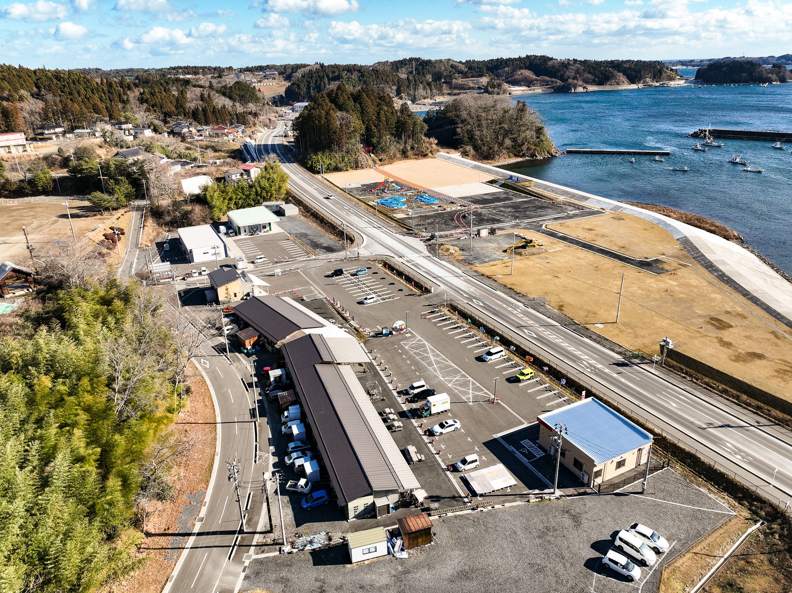 中央に曲がりくねった道路が走り、道の両側には駐車場と建物が立ち並び、その奥には広い埋め立て地と湾が広がっている海沿いの復興地域の風景を高台から定点カメラで撮影した写真