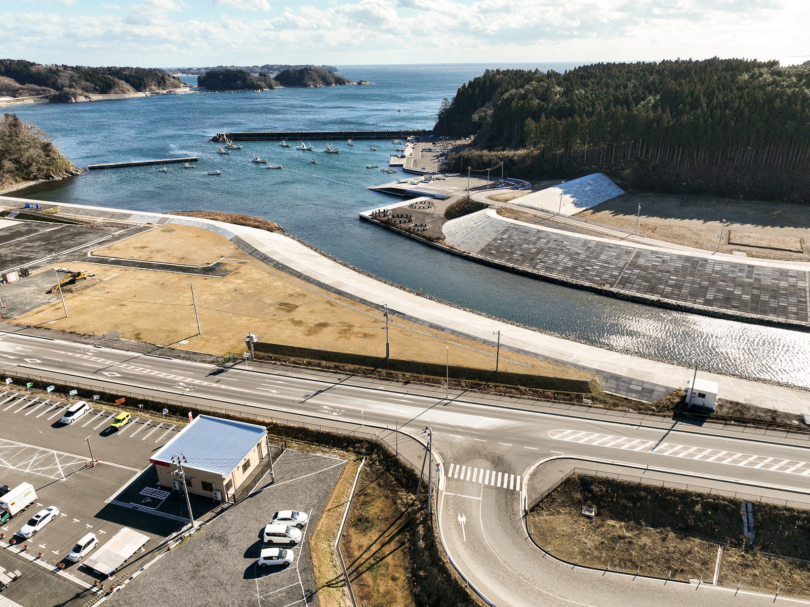 海に面した港湾地域で、防潮堤に囲まれた湾内には複数の漁船が浮かび、周囲には山と森林が広がっている防災工事中の風景を高台から定点カメラで撮影した写真