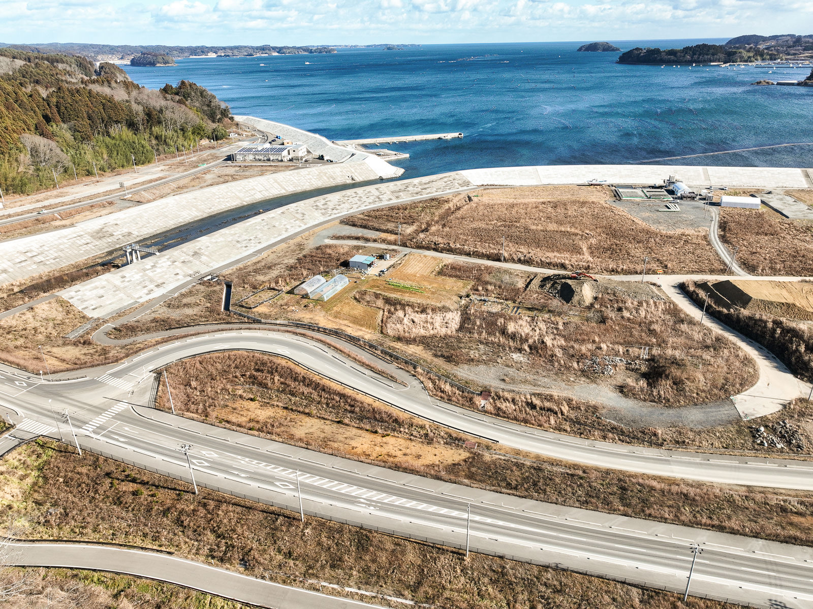 防潮堤が海岸線に沿って伸び、その手前には草地や農地の跡地と思われる区画と小さな建物が点在し、奥に太平洋が広がっている昼間の風景を高台から定点カメラで撮影した写真