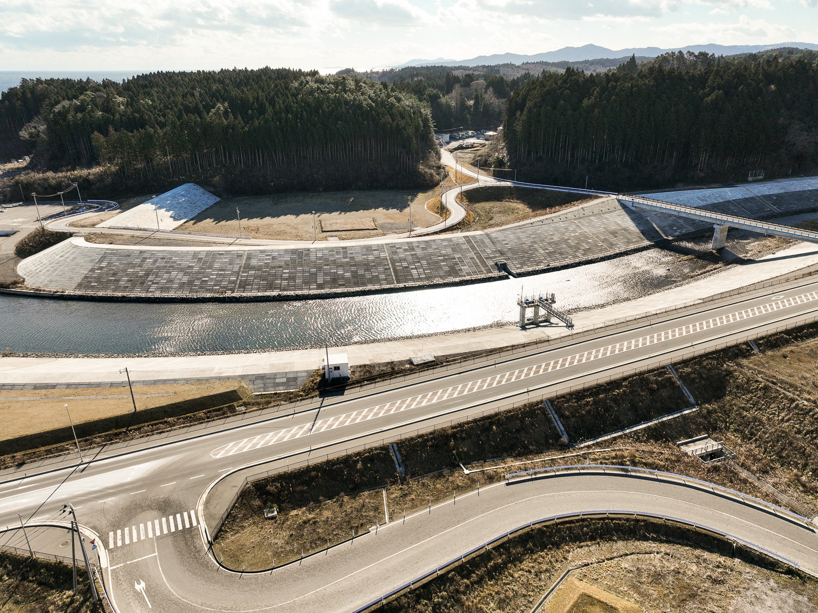 森に囲まれた川の上に橋が架かり、その両岸には整地され建設作業中の土地が広がる風景を高台から定点カメラで撮影した写真
