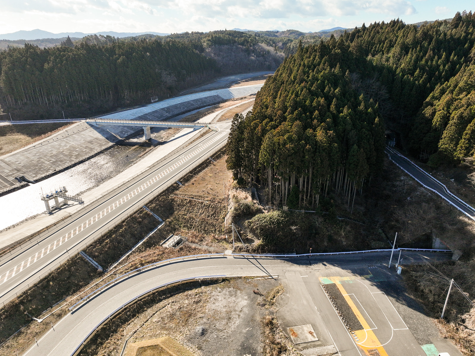 護岸工事が行われた川にかけられた橋が森の中に延び、両側に広がる山道と防潮堤が整然と整備された様子の風景を高台から定点カメラで撮影した写真