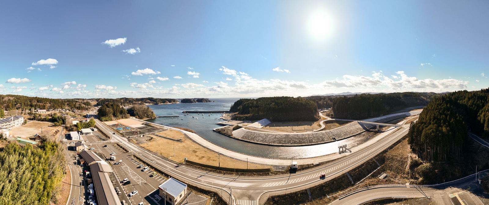 川が湾へ流れ込む地点に湾曲した白い堤防が広がり、右手に青緑色の針葉樹林が密集し、左手に小さな街並みと港が見える自然と人工物が共存する海岸の風景を高台から定点カメラで撮影した写真