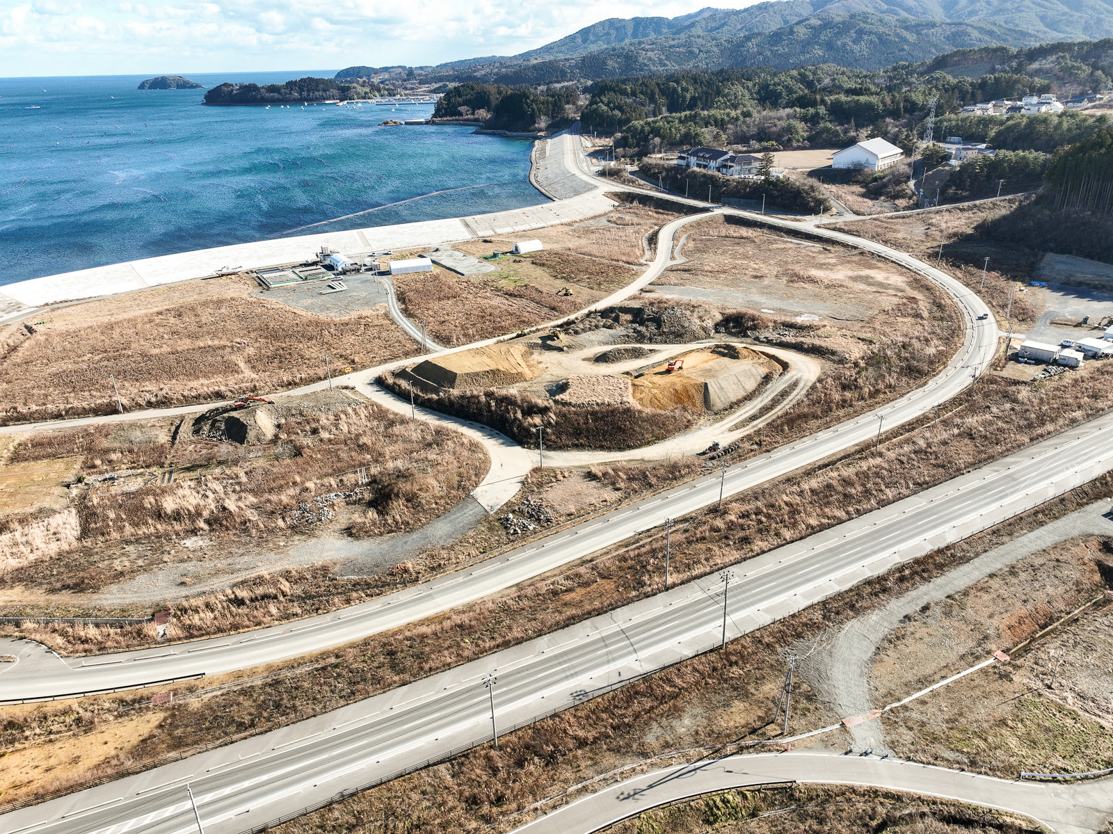 右手に湾の入口と山々が見え、中央には工事中の土地や造成された道、左奥には防潮堤に囲まれた海と岸が確認できる広域の復興作業地の昼間の風景を高台から定点カメラで撮影した写真
