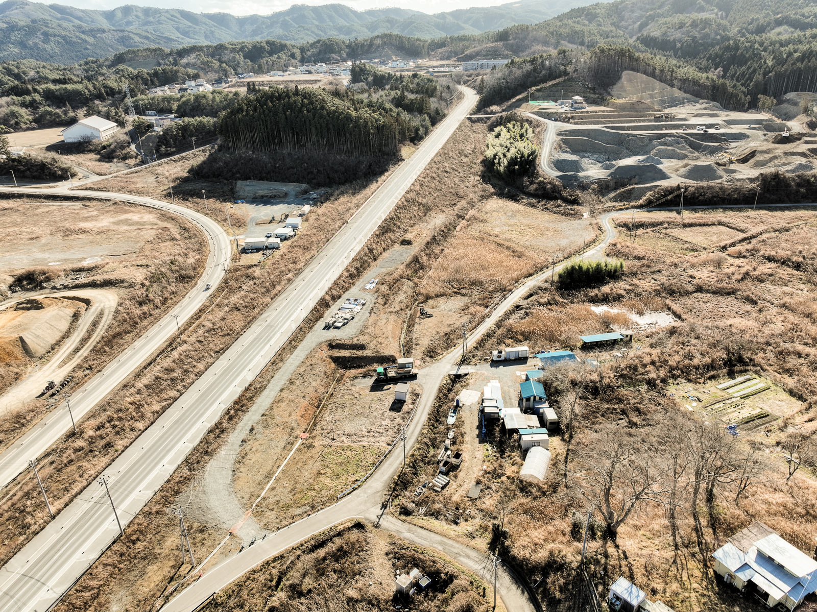 中央を縦に貫く広い道路の両側に造成中の土地や工事車両が点在し、奥には木々に囲まれた小高い丘と建物群、さらに背景に山々が連なる昼間の風景を高台から定点カメラで撮影した写真
