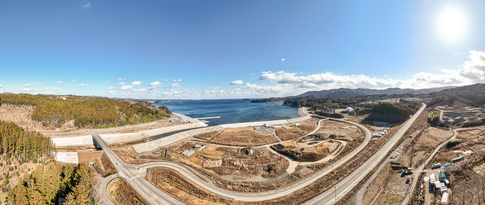 海に面した地域を広角で捉えており、手前に複雑に入り組んだ道路と高く盛られた防潮堤、奥には広がる海と空が見える広大な昼間の風景を高台から定点カメラで撮影した写真