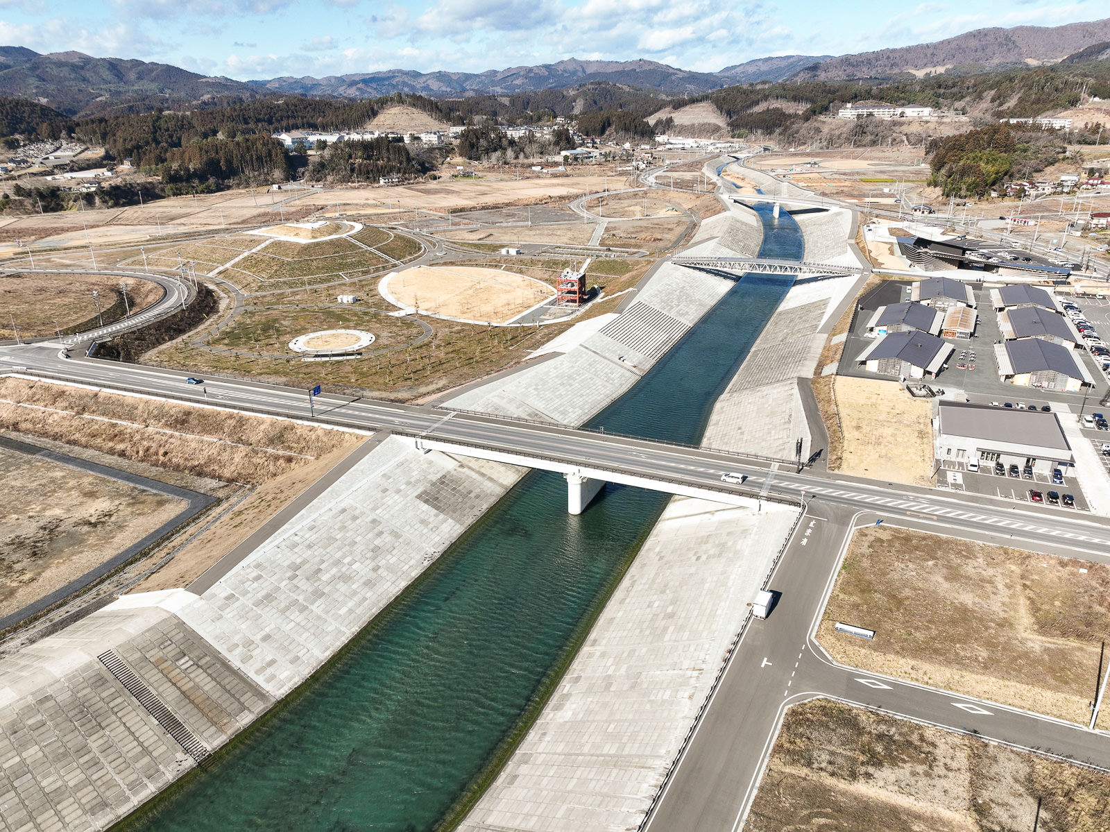 直線的に奥まで流れる川と高いコンクリート護岸、その周囲に円形や曲線の道路が巡る再整備された土地が広がり、背景には森と山が重なる昼間の風景を高台から定点カメラで撮影した写真