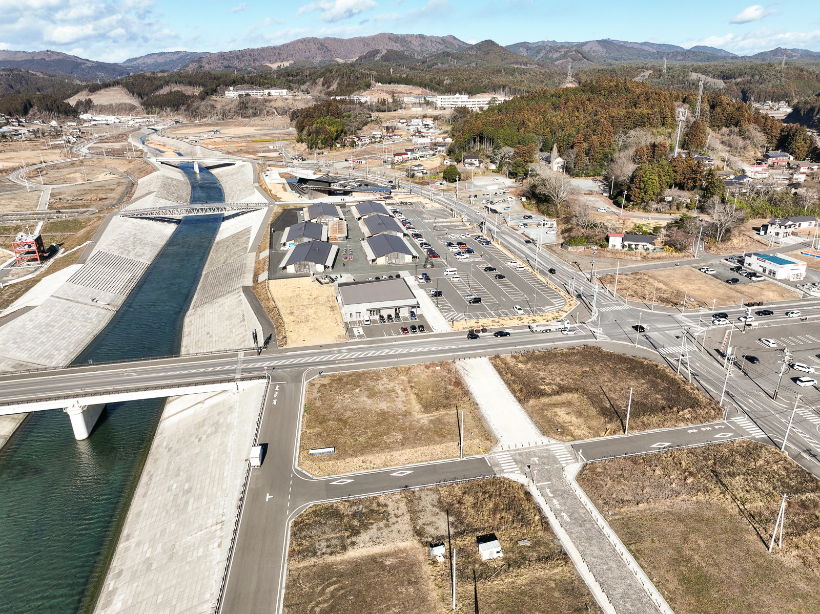 河川の護岸が高く整備され、川沿いに新しく建てられた建物群と広い駐車場が並び、手前には大きな道路と橋が川を横切っている様子の昼間の風景を高台から定点カメラで撮影した写真
