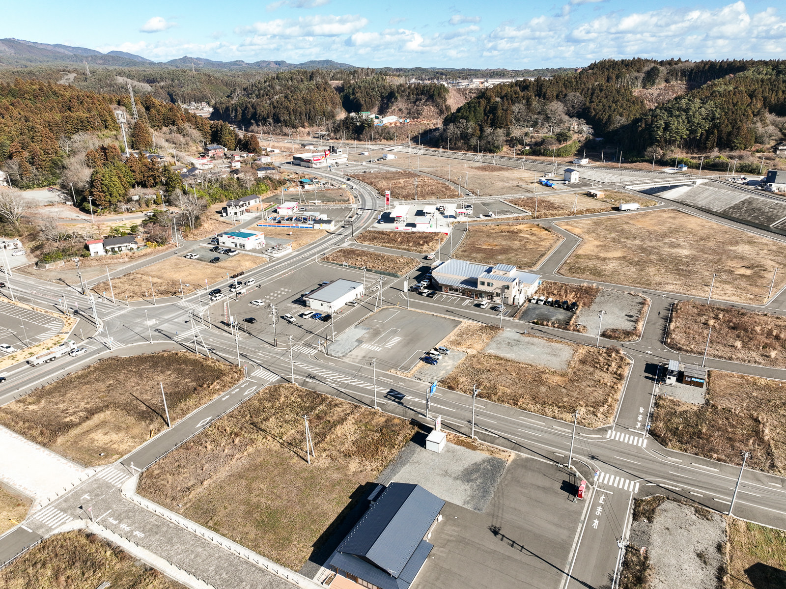 交差点を中心に区画整理された土地と建物が整然と並び、背景には山林が広がっている復興中の市街地の昼間の風景を高台から定点カメラで撮影した写真