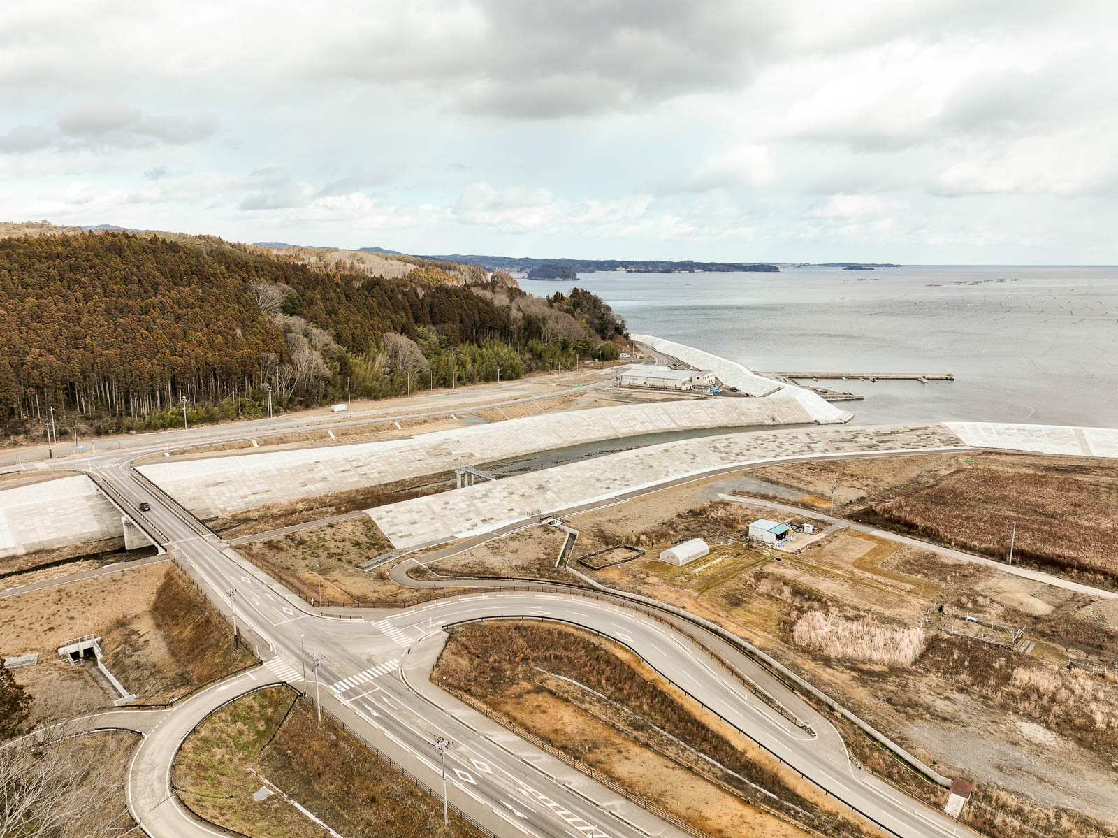 海に近いエリアで山林と海の間に長く延びた防潮堤と交差する道路が確認でき、全体的に復興工事中の様子がうかがえる昼間の風景を高台から定点カメラで撮影した写真