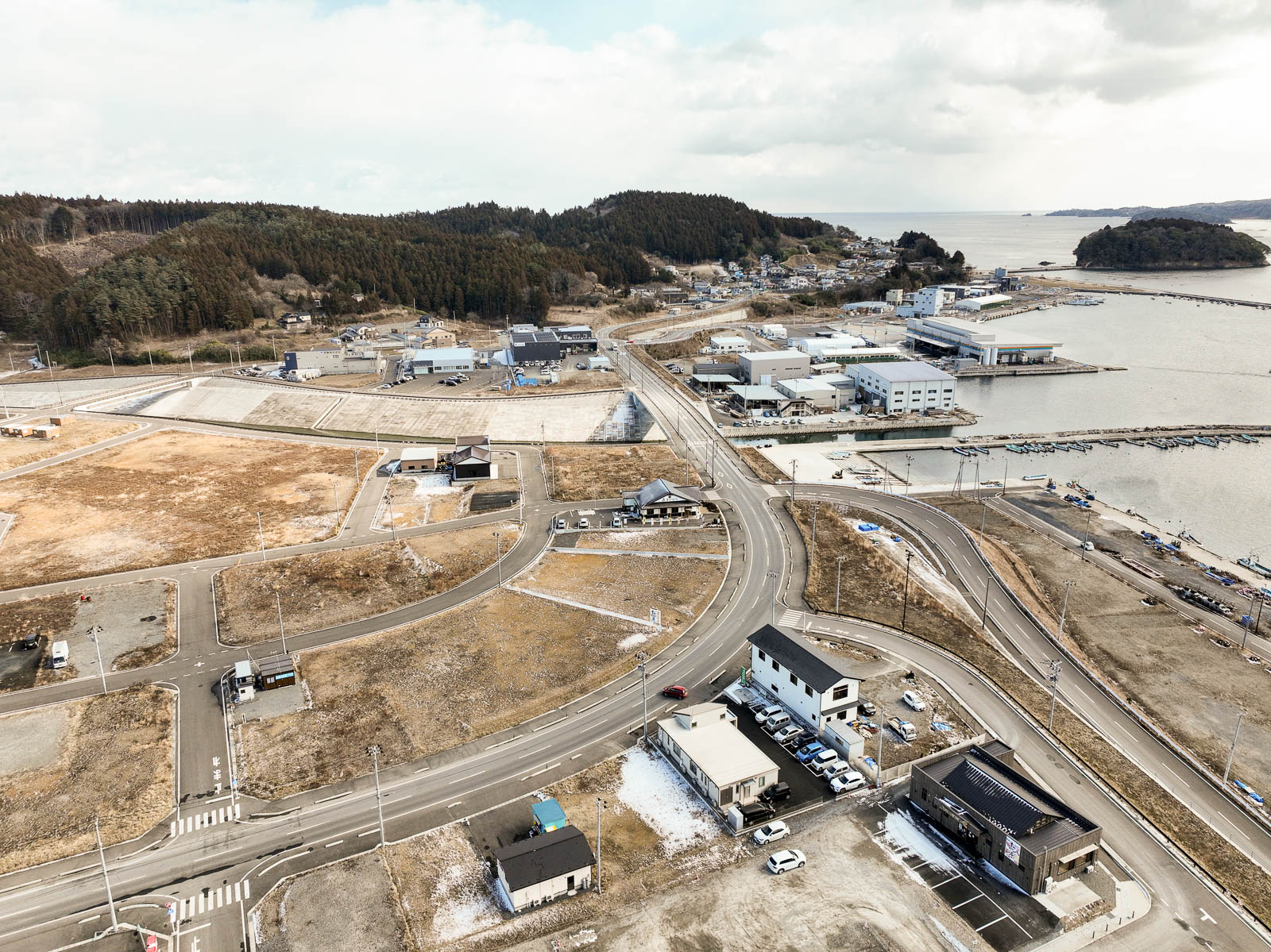 道路が交差し、住宅や建物がまばらに建ち並び、奥には森林が広がっている海の近くにある小さな町の風景を高台から定点カメラで撮影した写真