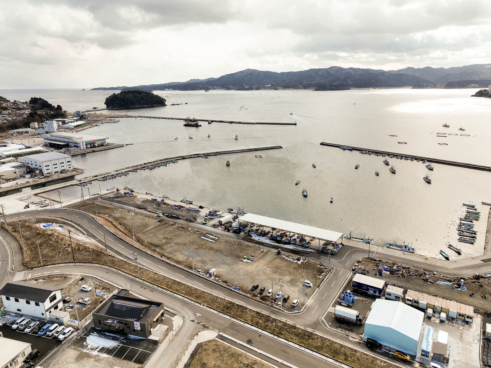 防波堤内に漁船が浮かび、岸には白い倉庫や工場が整然と並んでいる海に面した漁港の風景を高台から定点カメラで撮影した写真