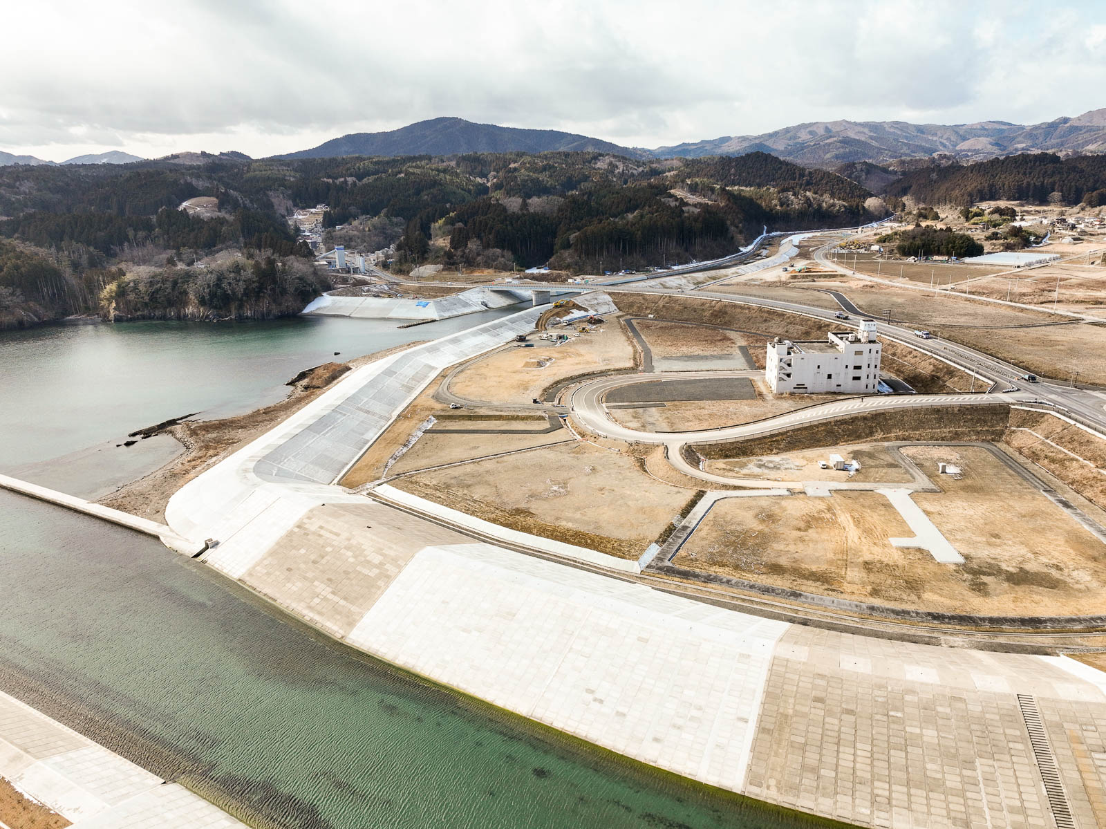 海へ流れ込む川の両岸に高さのある堤防が築かれ、その奥には曲線を描く道路と、白い避難ビルのような建物が広がる造成地とともに、山に囲まれた静かな風景を高台から定点カメラで撮影した写真