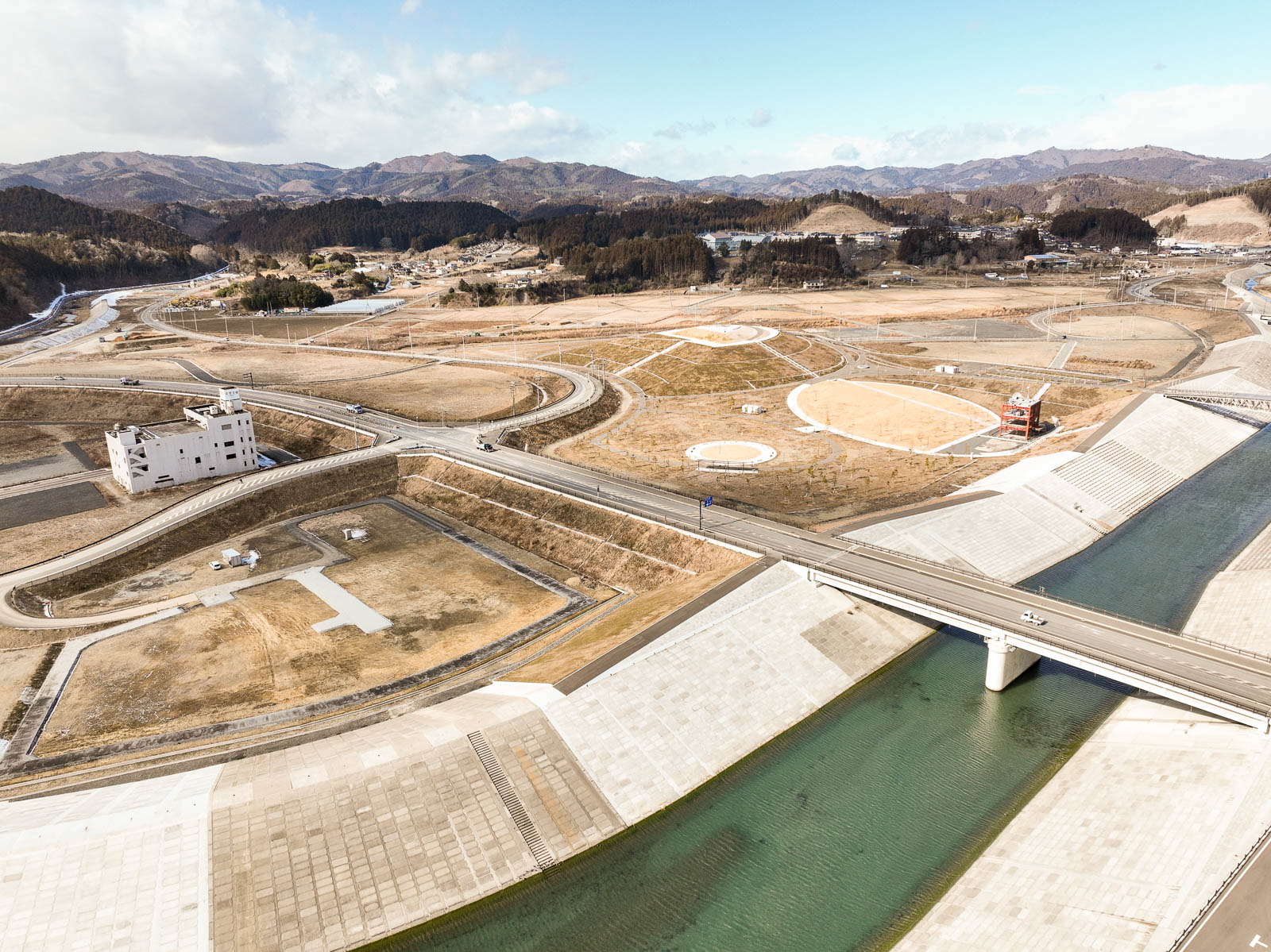 川の上にかかる大きな橋を中心に、その奥には円形の公園や広場、赤い建造物が点在し、さらに背後には山々が広がる整備された土地が一望できる風景を高台から定点カメラで撮影した写真