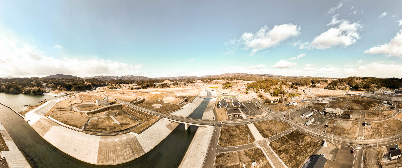 手前に川と防潮堤、中央に大きな橋と新しく整備された道路網、その奥に広がる山々と街並みがパノラマで写されており、復興が進んだ都市計画の昼間の風景を高台から定点カメラで撮影した写真