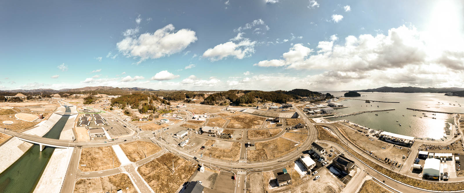 手前から奥へと川と橋が続き、右手には広く整備された街並みと道路網、さらにその先に港と海が広がっており、港湾地域の再整備の様子の昼間の風景を高台から定点カメラで撮影した写真