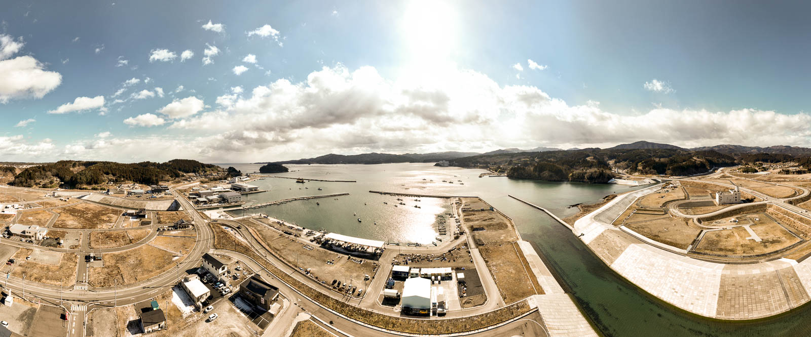 中央に太陽が輝き、左右に湾と堤防、周囲に山々と整備された土地が広がっている湾に面した町の風景を高台から定点カメラで撮影した写真
