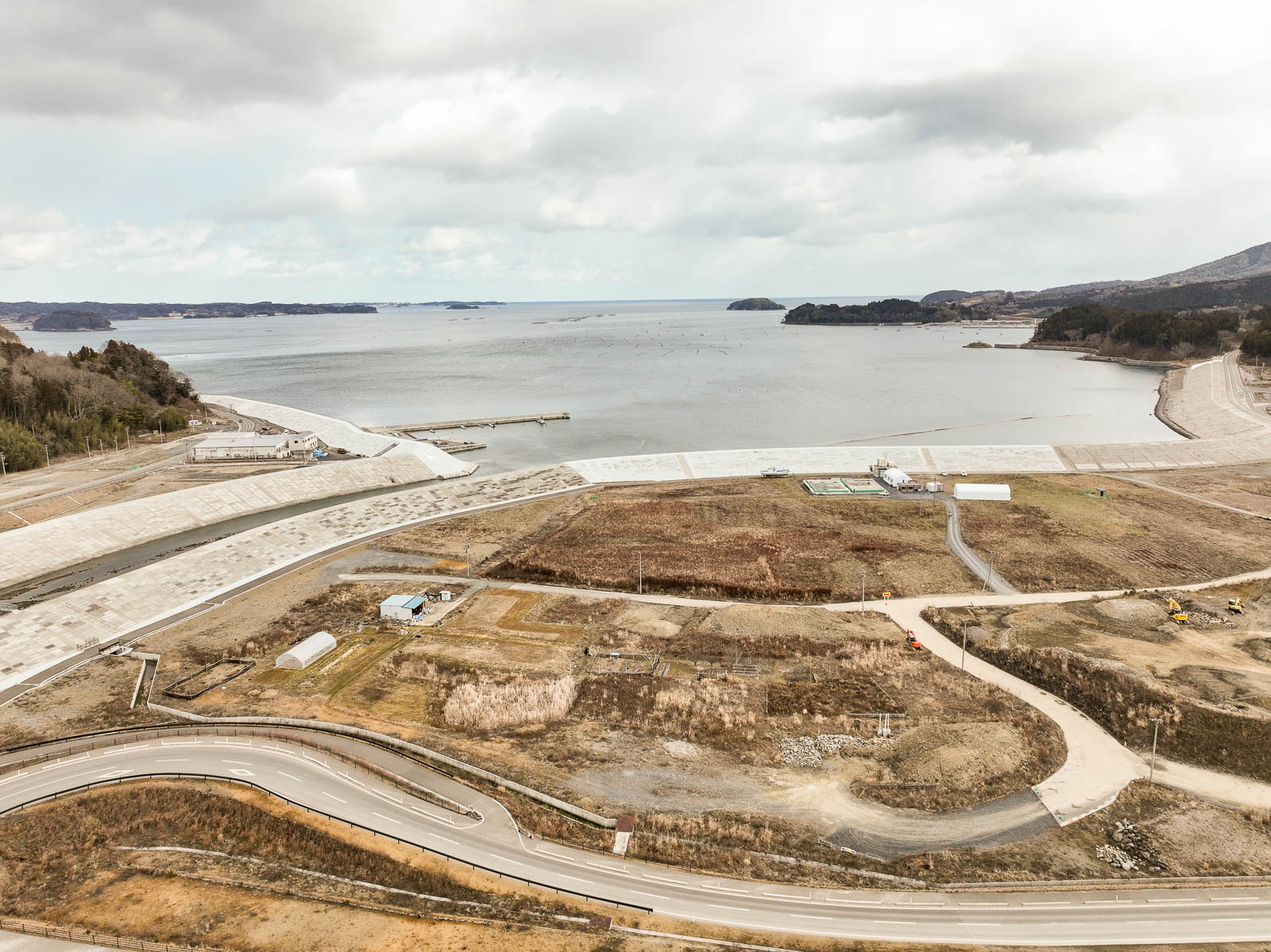防潮堤が海岸線に沿って伸び、その手前には草地や農地の跡地と思われる区画と小さな建物が点在し、奥に太平洋が広がっている昼間の風景を高台から定点カメラで撮影した写真