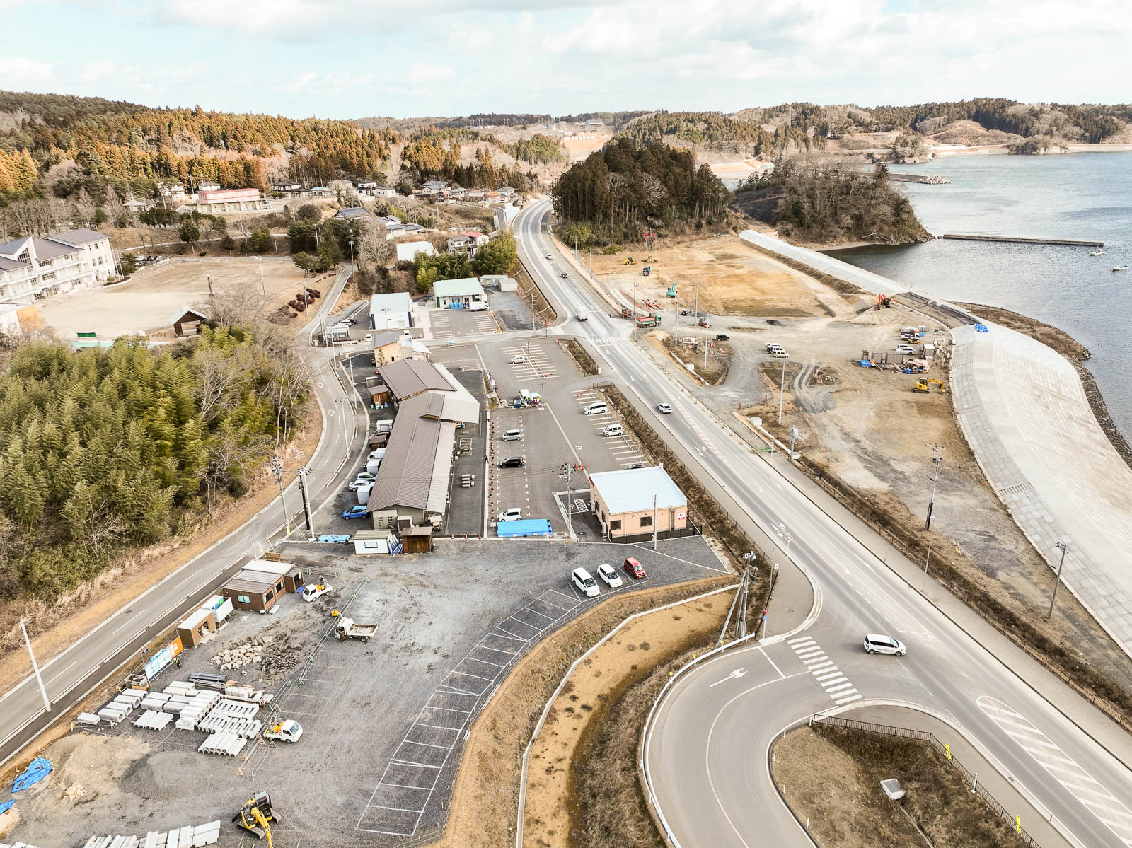 中央に曲がりくねった道路が走り、道の両側には駐車場と建物が立ち並び、その奥には広い埋め立て地と湾が広がっている海沿いの復興地域の風景を高台から定点カメラで撮影した写真