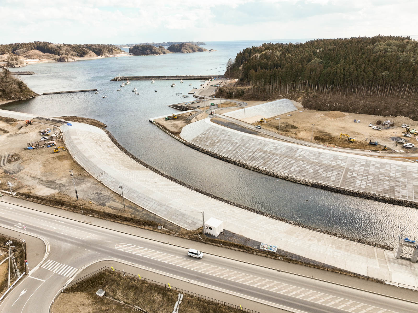 海に面した港湾地域で、防潮堤に囲まれた湾内には複数の漁船が浮かび、周囲には山と森林が広がっている防災工事中の風景を高台から定点カメラで撮影した写真