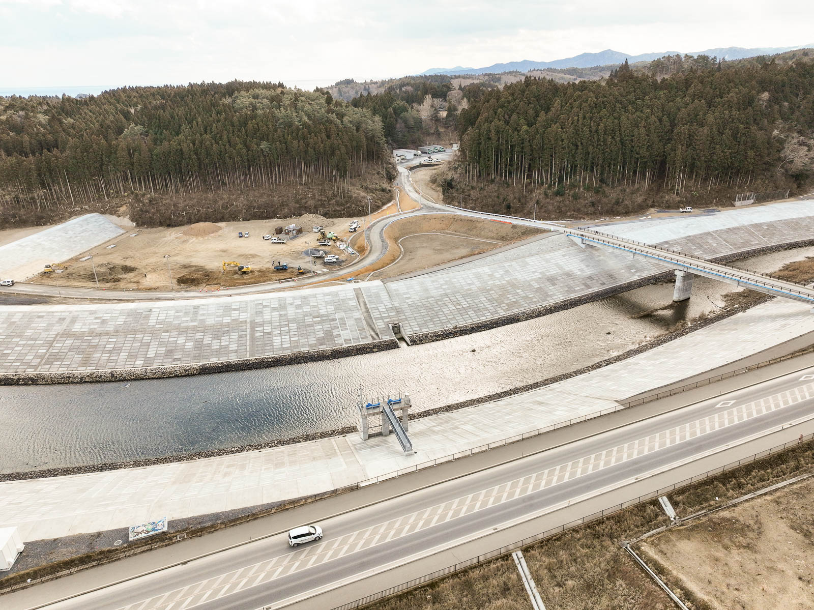森に囲まれた川の上に橋が架かり、その両岸には整地され建設作業中の土地が広がる風景を高台から定点カメラで撮影した写真