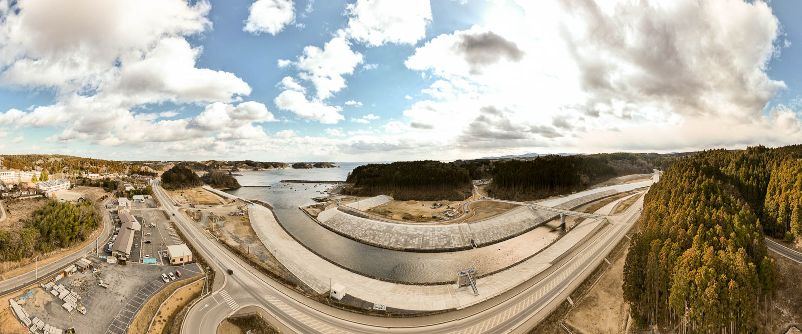 川が湾へ流れ込む地点に湾曲した白い堤防が広がり、右手に青緑色の針葉樹林が密集し、左手に小さな街並みと港が見える自然と人工物が共存する海岸の風景を高台から定点カメラで撮影した写真