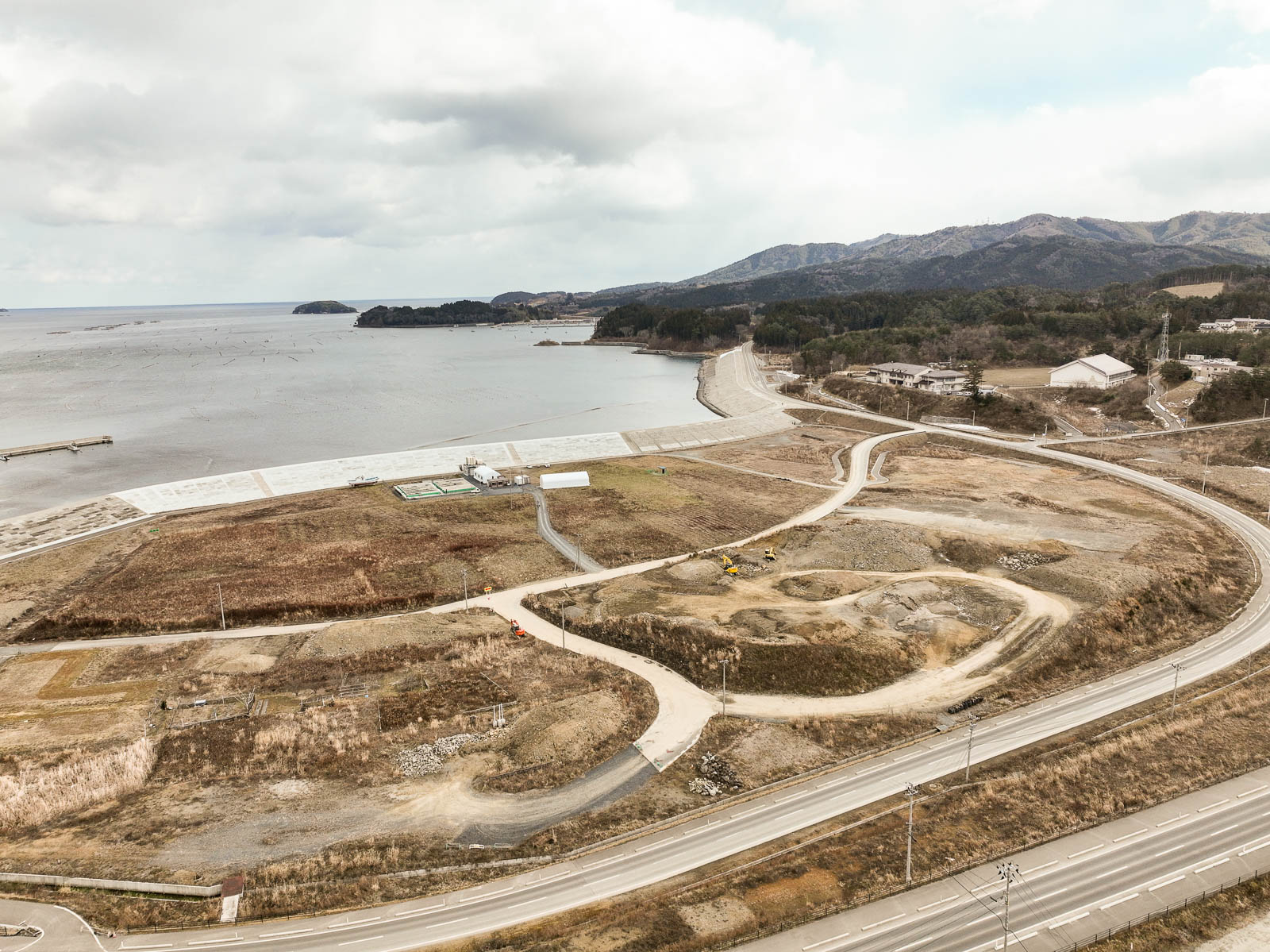 右手に湾の入口と山々が見え、中央には工事中の土地や造成された道、左奥には防潮堤に囲まれた海と岸が確認できる広域の復興作業地の昼間の風景を高台から定点カメラで撮影した写真