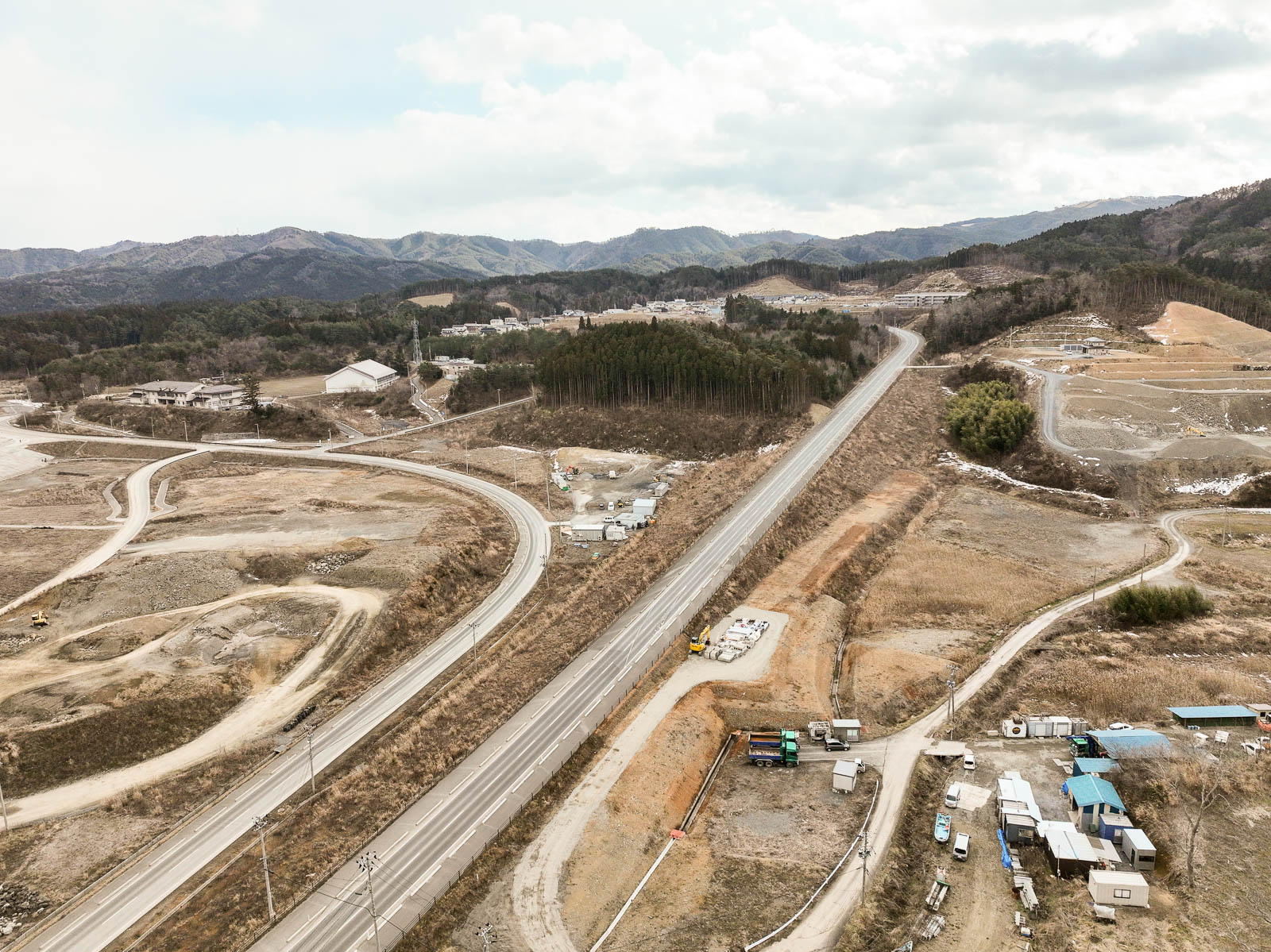 中央を縦に貫く広い道路の両側に造成中の土地や工事車両が点在し、奥には木々に囲まれた小高い丘と建物群、さらに背景に山々が連なる昼間の風景を高台から定点カメラで撮影した写真