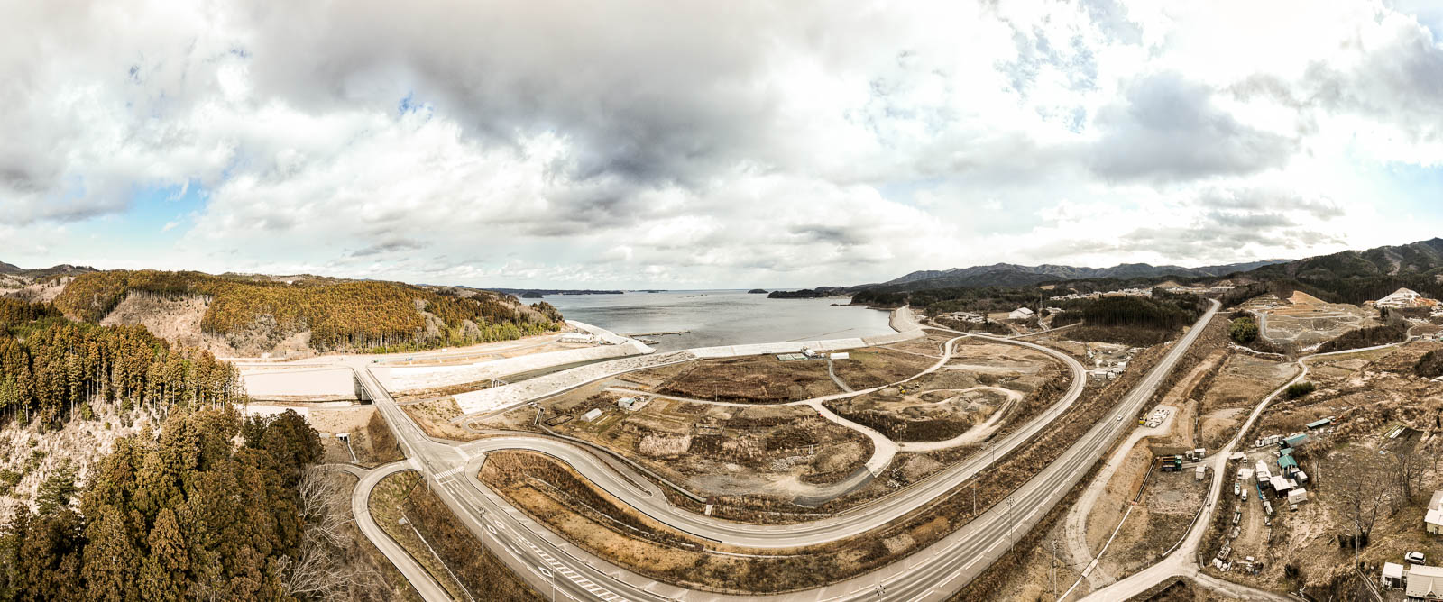 海に面した地域を広角で捉えており、手前に複雑に入り組んだ道路と高く盛られた防潮堤、奥には広がる海と空が見える広大な昼間の風景を高台から定点カメラで撮影した写真