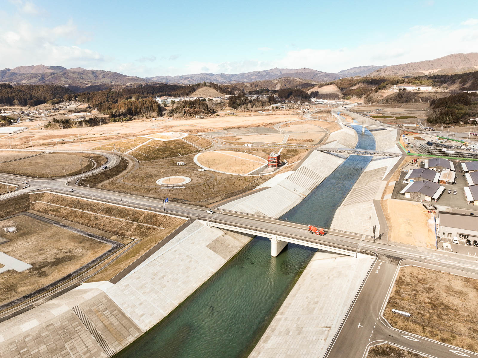 直線的に奥まで流れる川と高いコンクリート護岸、その周囲に円形や曲線の道路が巡る再整備された土地が広がり、背景には森と山が重なる昼間の風景を高台から定点カメラで撮影した写真