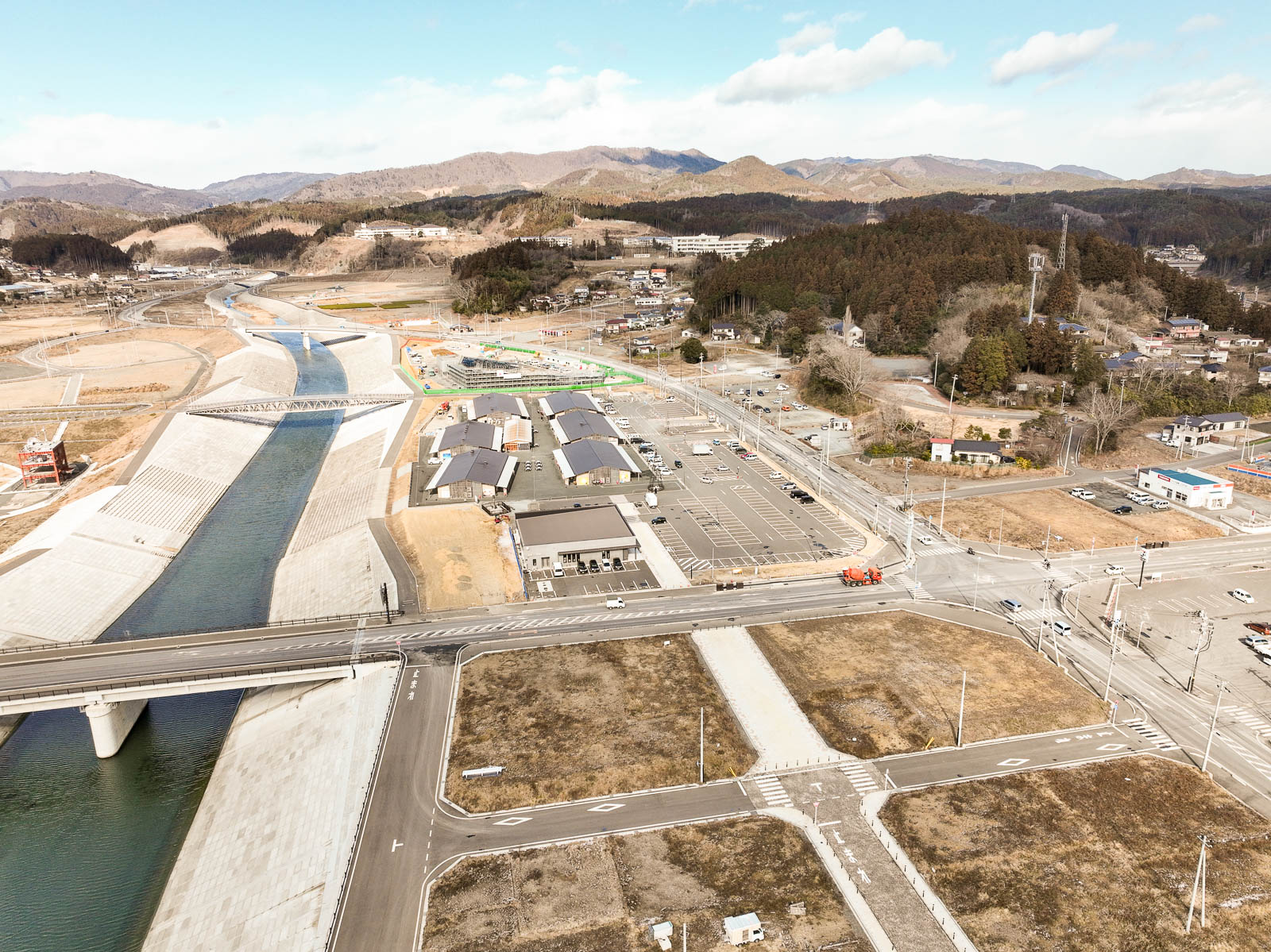 河川の護岸が高く整備され、川沿いに新しく建てられた建物群と広い駐車場が並び、手前には大きな道路と橋が川を横切っている様子の昼間の風景を高台から定点カメラで撮影した写真