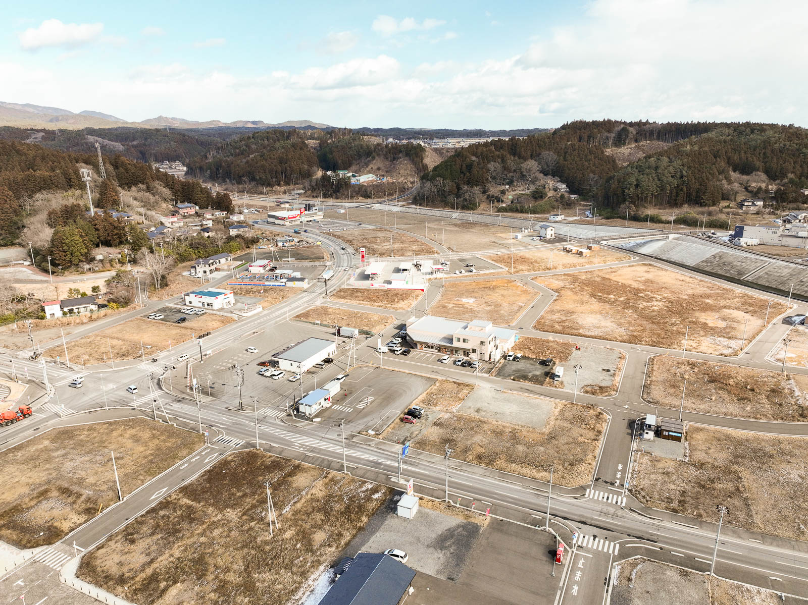 交差点を中心に区画整理された土地と建物が整然と並び、背景には山林が広がっている復興中の市街地の昼間の風景を高台から定点カメラで撮影した写真