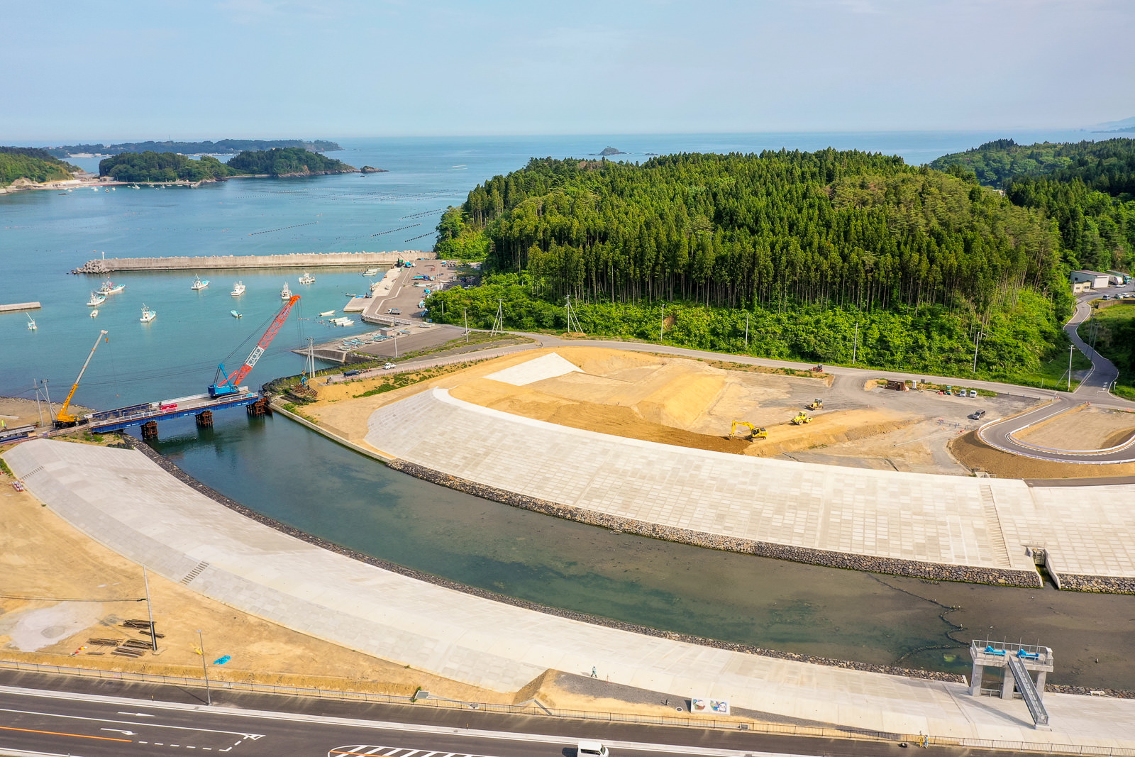 川のような水域が流れる沿岸の工事現場で、複数の建設車両や機材が配置され、背景には密集した森林が広がり、港には船とクレーンがあり、画像下部には道路が通っている写真