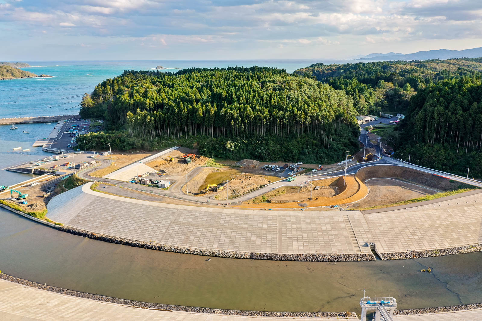 中央に広い工事現場と曲がりくねった道路、奥に港と防波堤、さらにその奥に海と空が見える。整備中の道路と防災施設の写真
