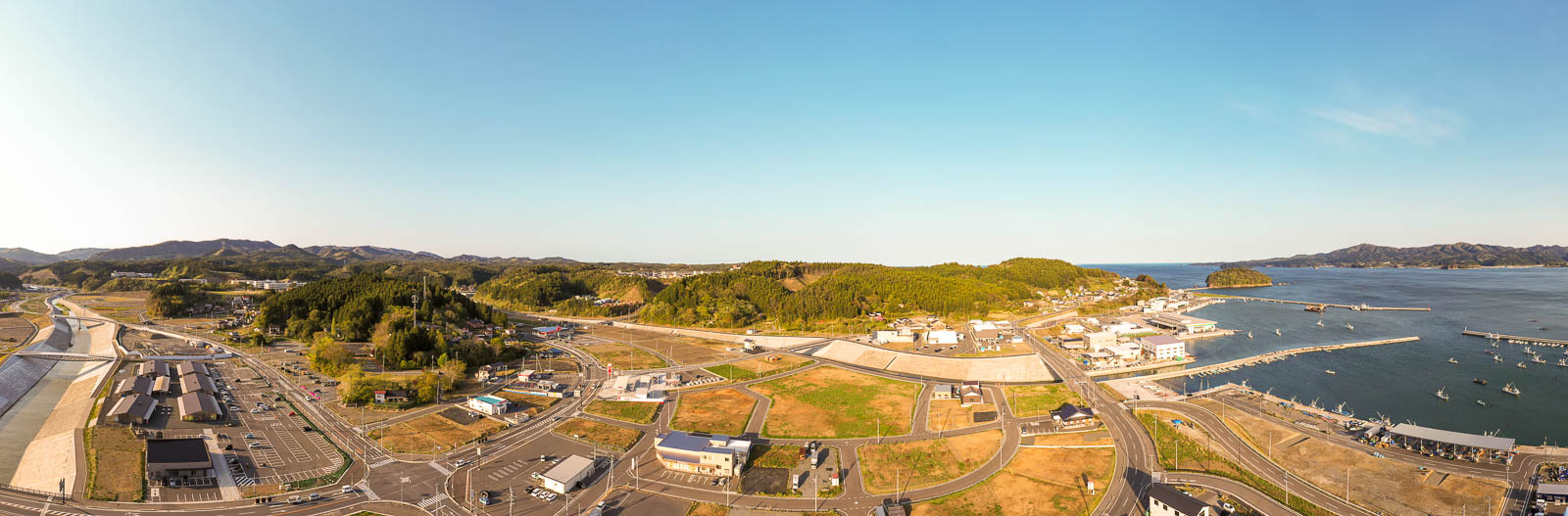左側には駐車場が見え、その横を堤防で整備された川が流れ中央部分には緑豊かな小高い丘があり、その背後には住宅地が広がっている写真