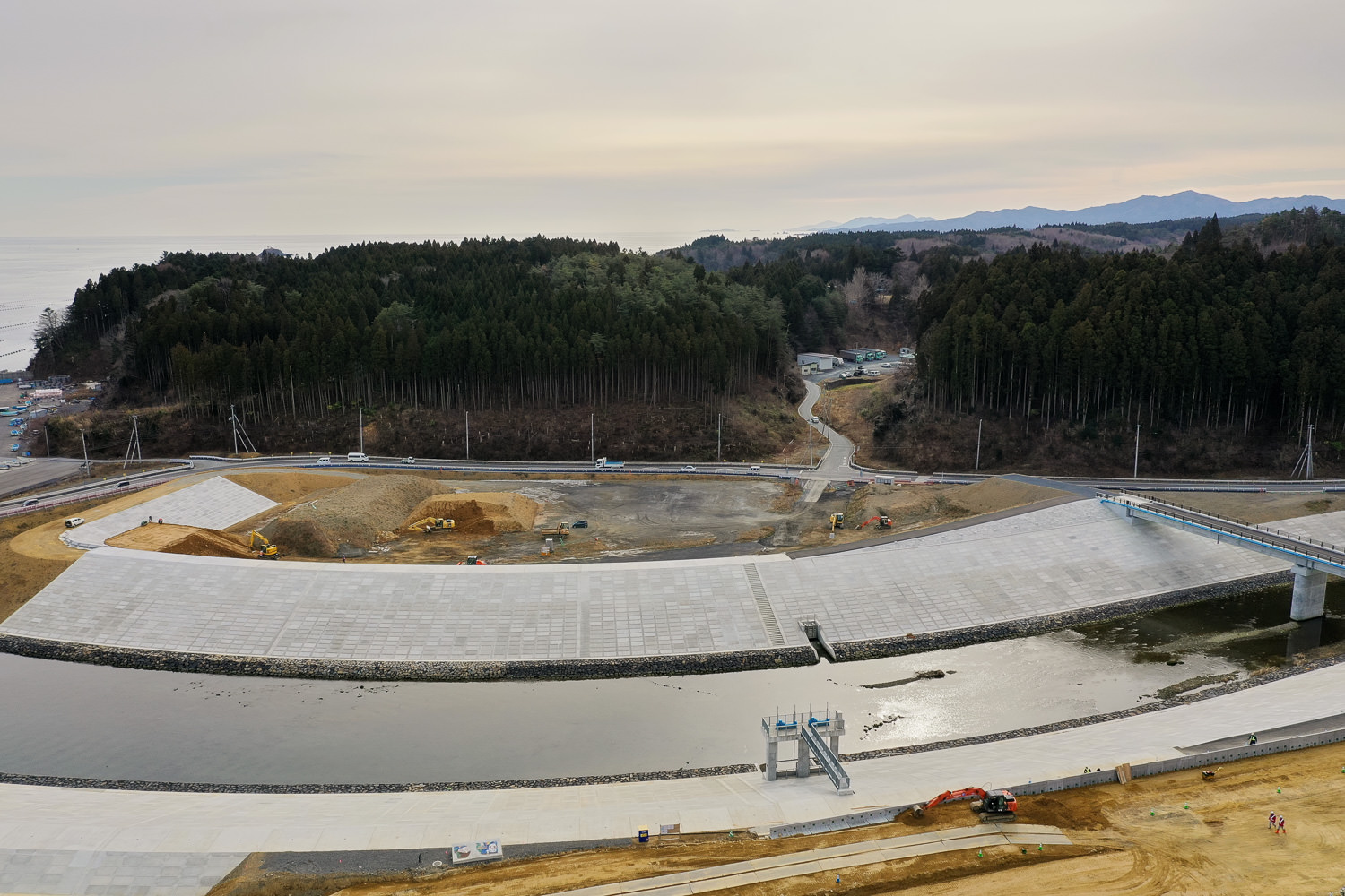 広いコンクリートの護岸が川の両側を囲み、背景には緑豊かな木々が茂る山とそのふもとを通る道路が見える写真