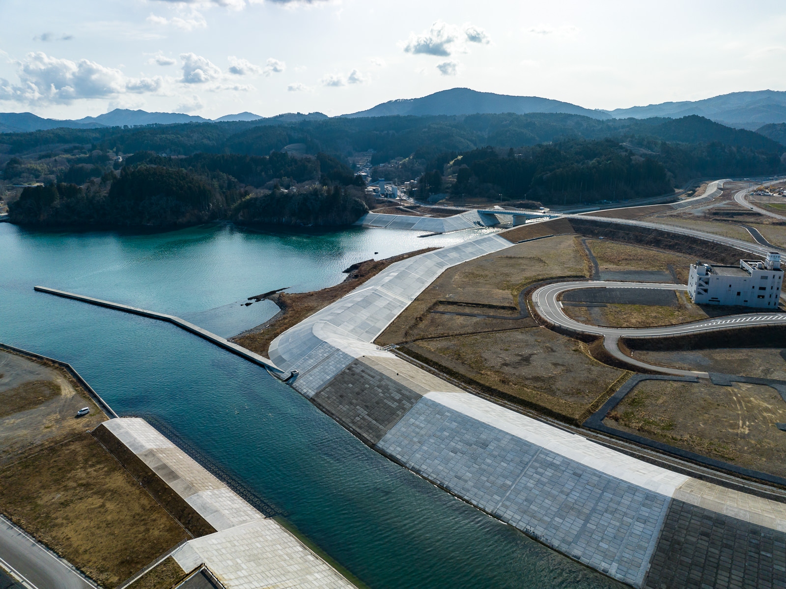 手前に広い川と白く整備された護岸が見え、奥には少し曲がった防波堤と海がつながっており、右手には新しく舗装された道路と白い建物があり、背景には山が重なる川の河口付近と防波堤の昼間の風景を高台から定点カメラで撮影した写真