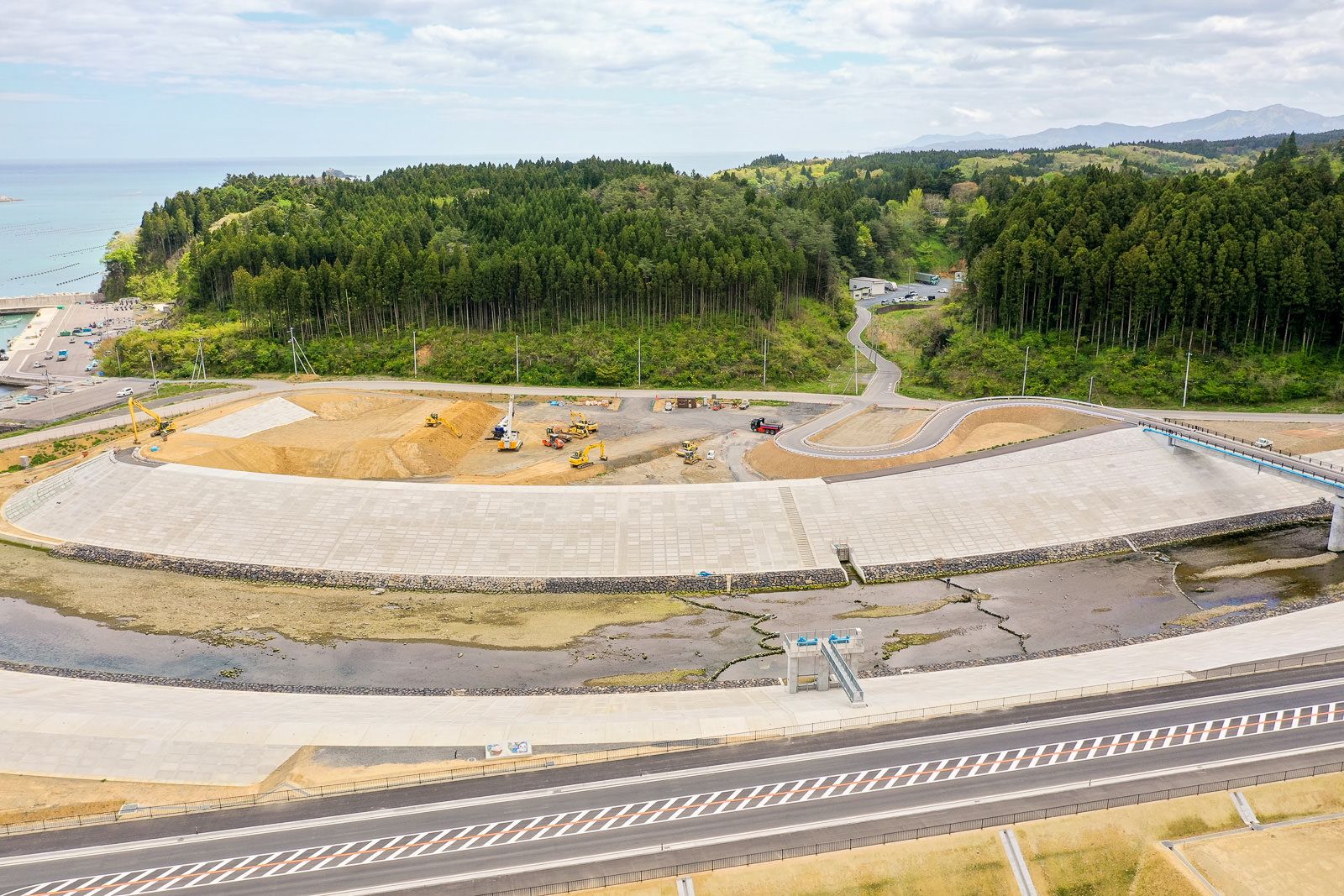 海岸沿いの工事現場で、手前に道路が走り、中央には川が流れ、その奥に重機や資材が並ぶ広いコンクリートの敷地と森が広がっている写真