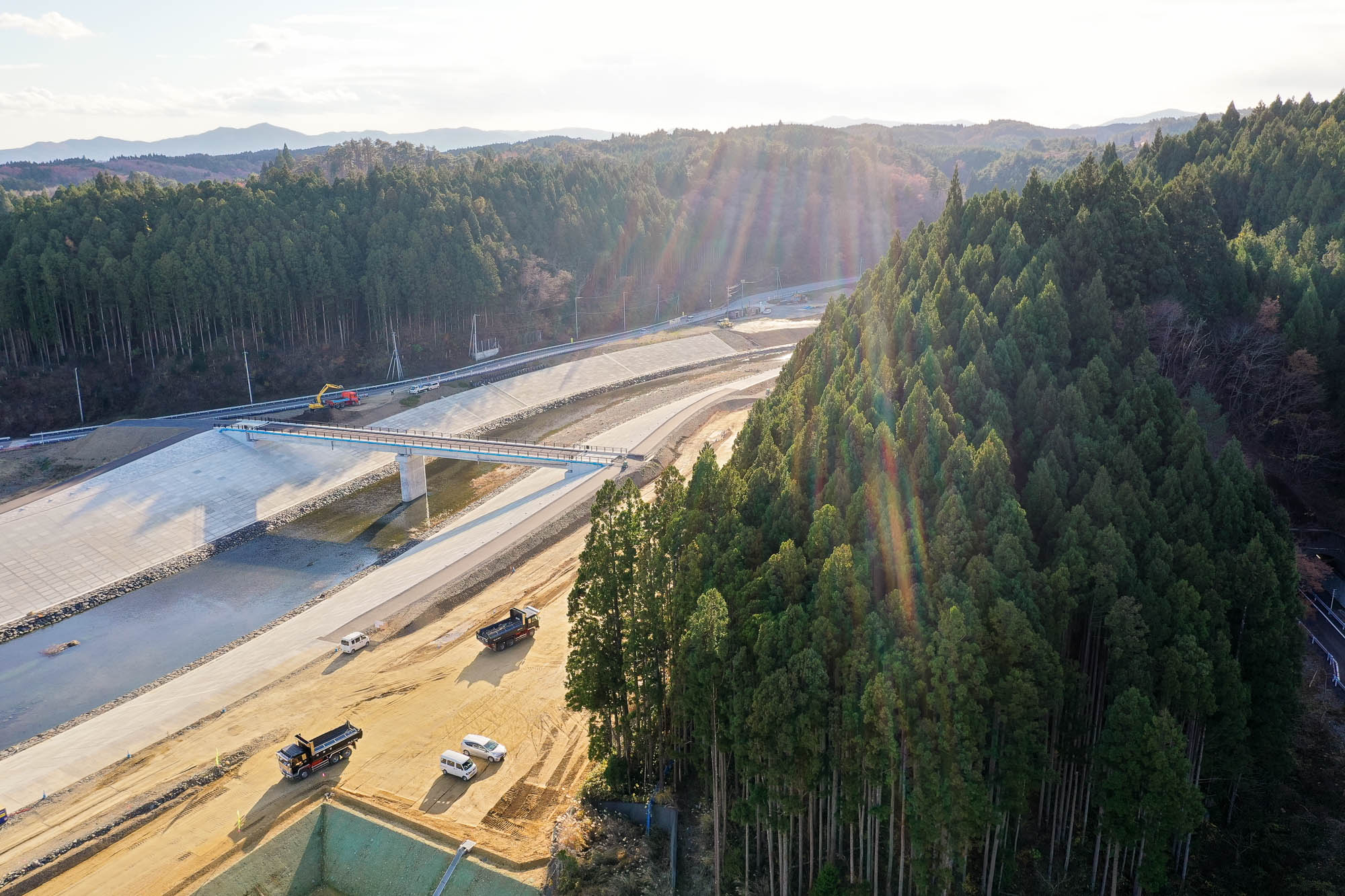 手前に工事中の道路と作業車両があり、右側に濃い緑の森林が広がり、左奥には川と橋が森林の中へ続いている様子の写真